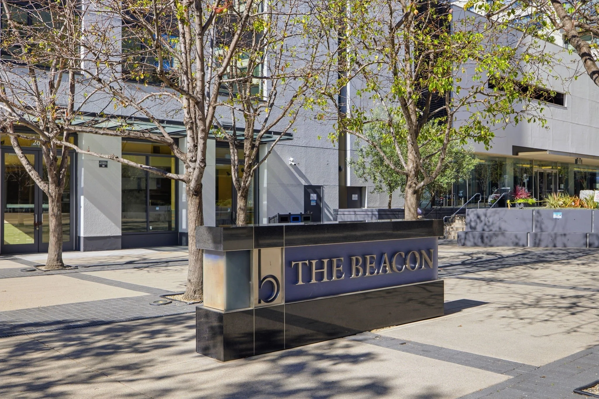 Street view of a modern commercial building with a black sign that reads 'The Beacon' and a large numeral 5. There are leafless trees along the sidewalk, willows or similar, with shadows cast on the ground. The building has large glass windows and a 