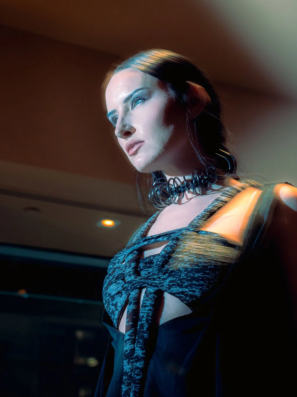 A woman with dark hair styled straight, wearing a black and metallic-patterned top, a black spiral choker necklace, and metallic jewelry, is looking off into the distance with a serious expression, illuminated by dramatic lighting in a dim indoor set