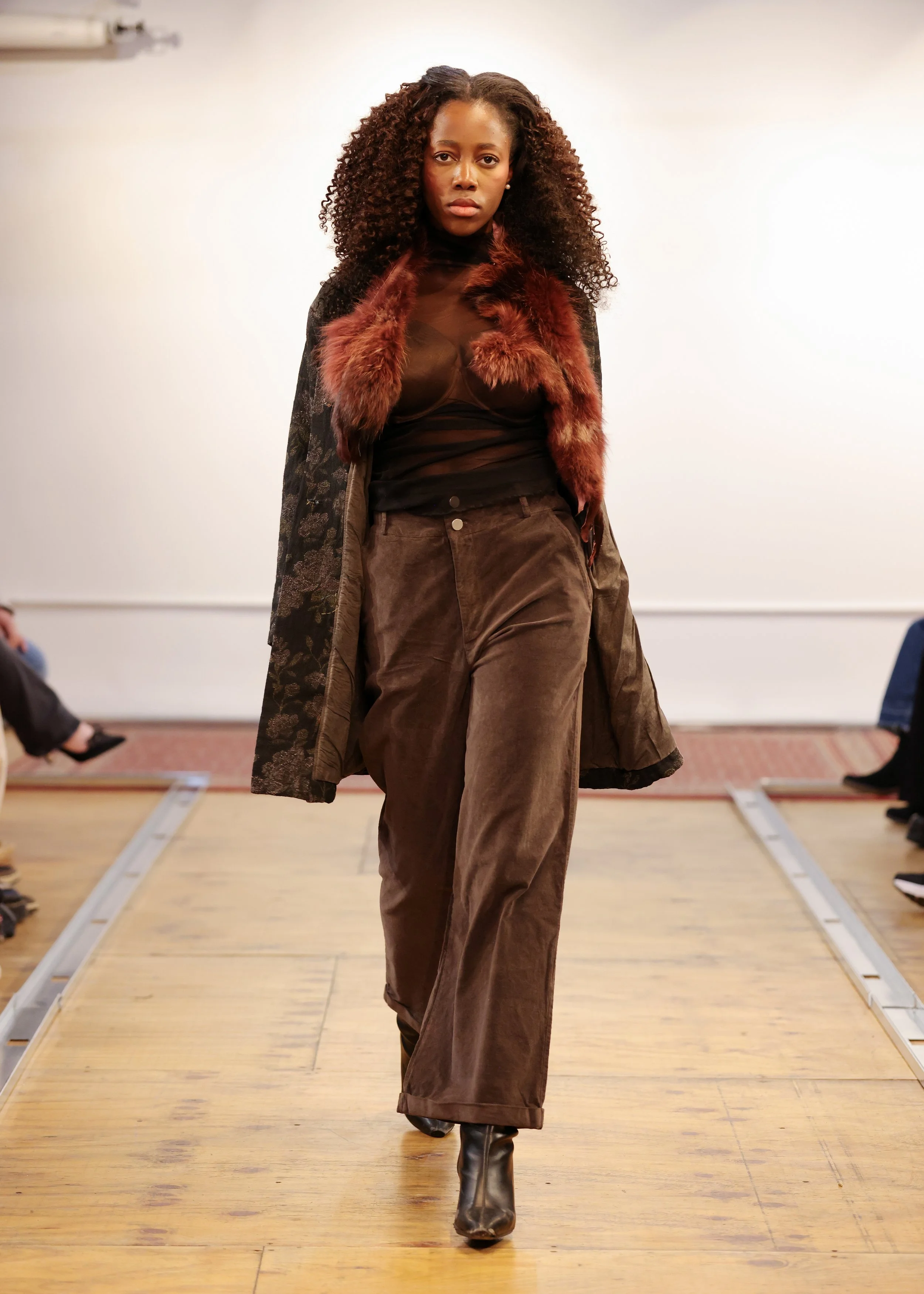 A woman walking down a runway during a fashion show, wearing brown wide-leg pants, a sheer black top, a patterned coat, and a fur stole.