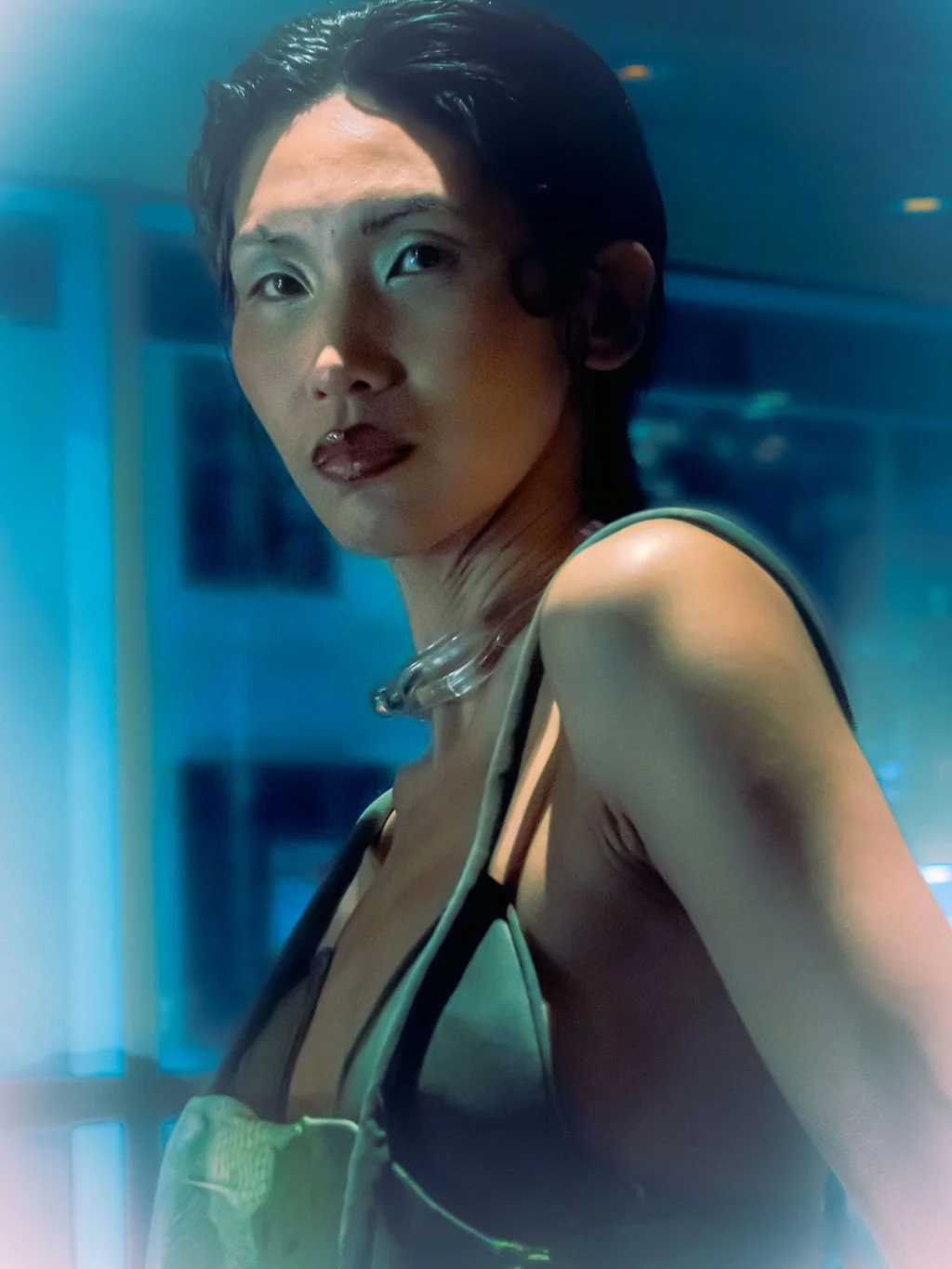 A woman with short, dark hair and dark lipstick looks over her shoulder in a dimly lit, futuristic setting with blue lighting.