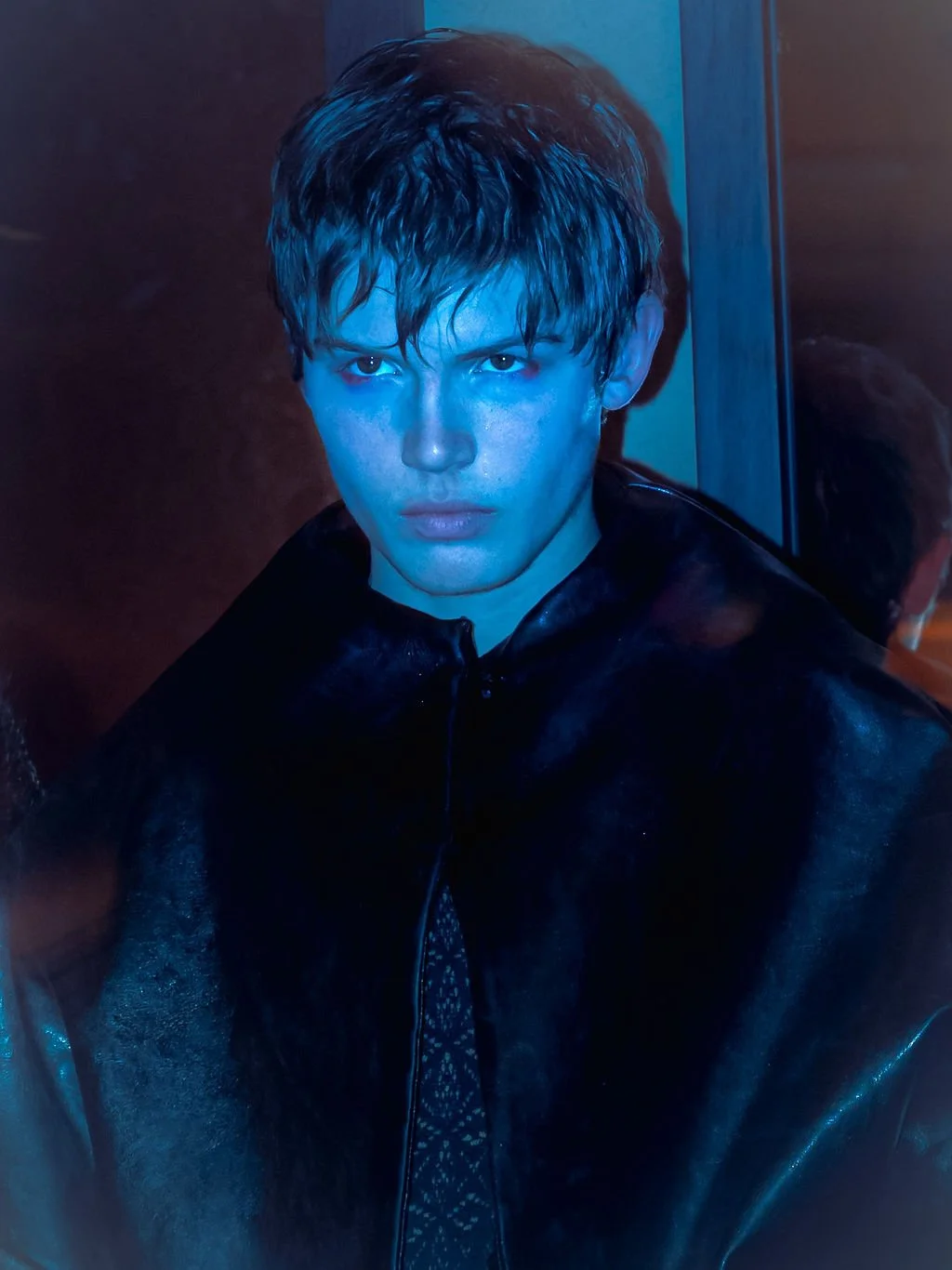 A young man with wet hair and a serious expression, wearing a shiny black leather jacket, standing in front of a mirror with blue lighting.
