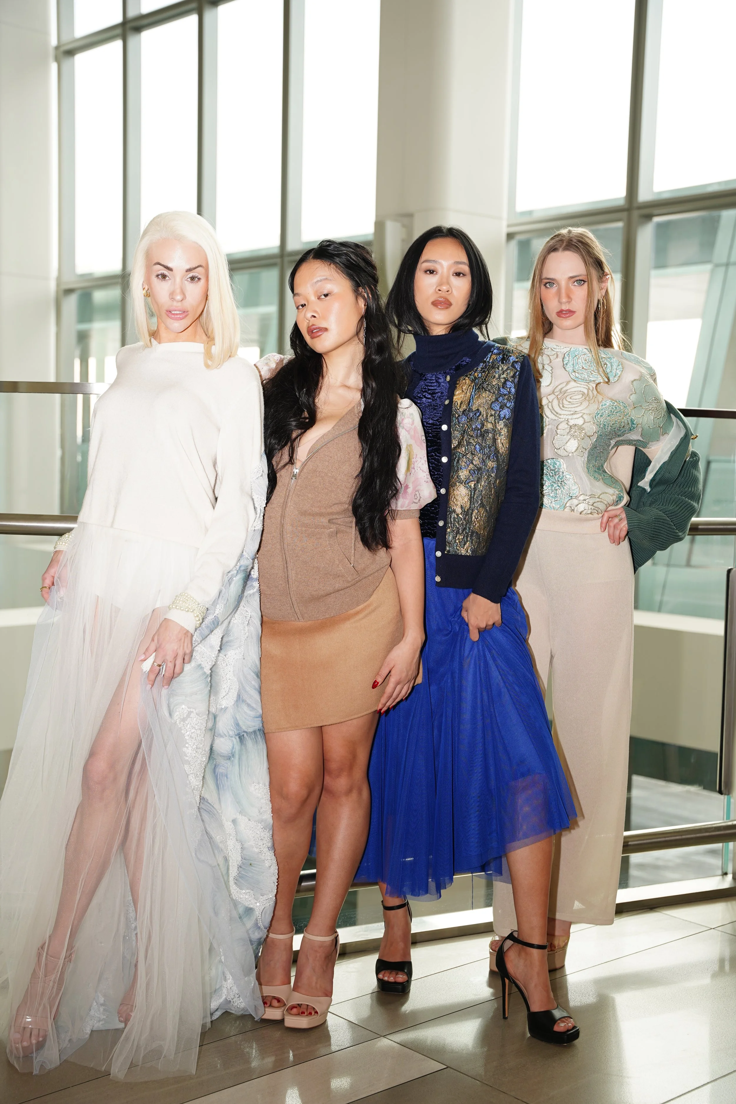 Four women standing together indoors, dressed in fashion-forward outfits, posing for a photo in front of large glass windows.
