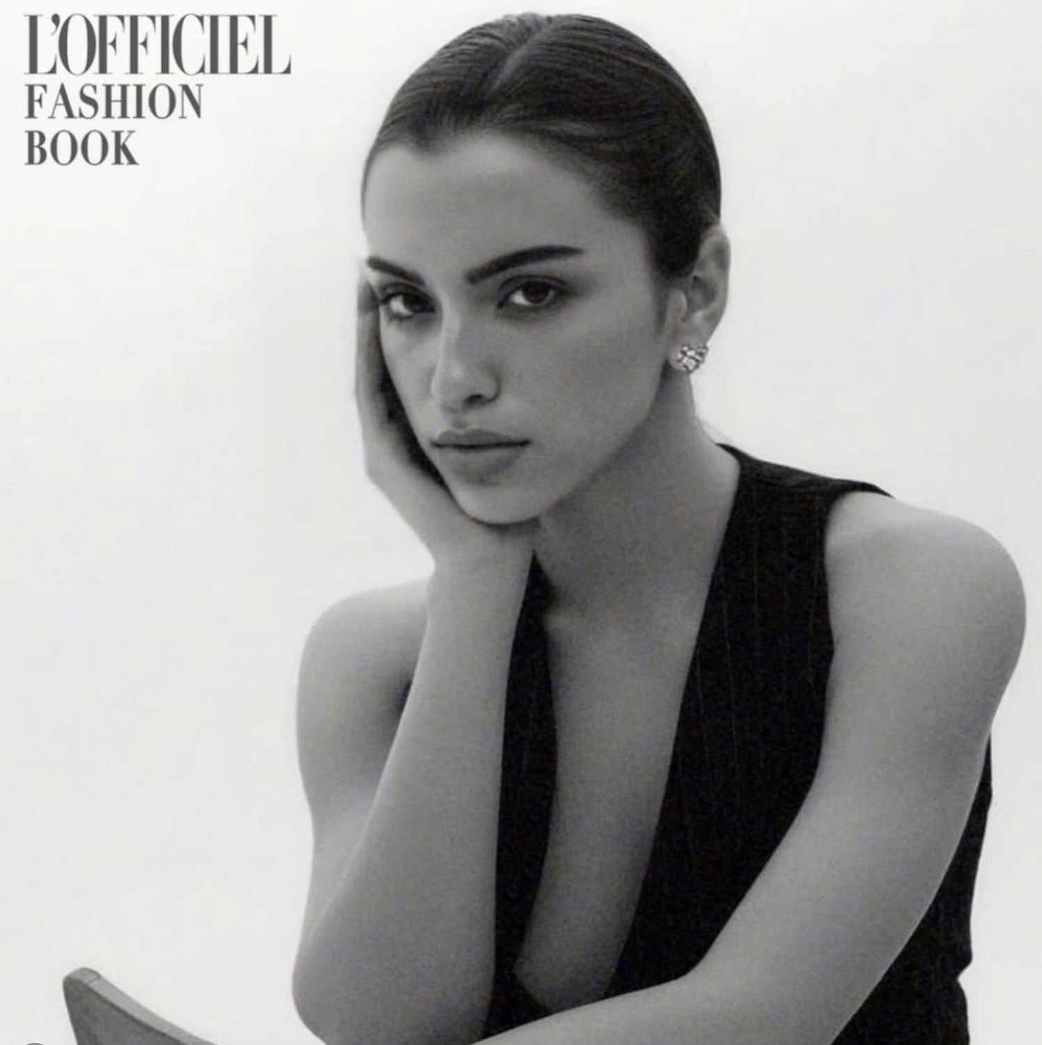 Black and white cover of L'Officiel Fashion Book featuring a woman with sleek dark hair, arched eyebrows, holding her face with her hand, wearing earrings and a sleeveless top.