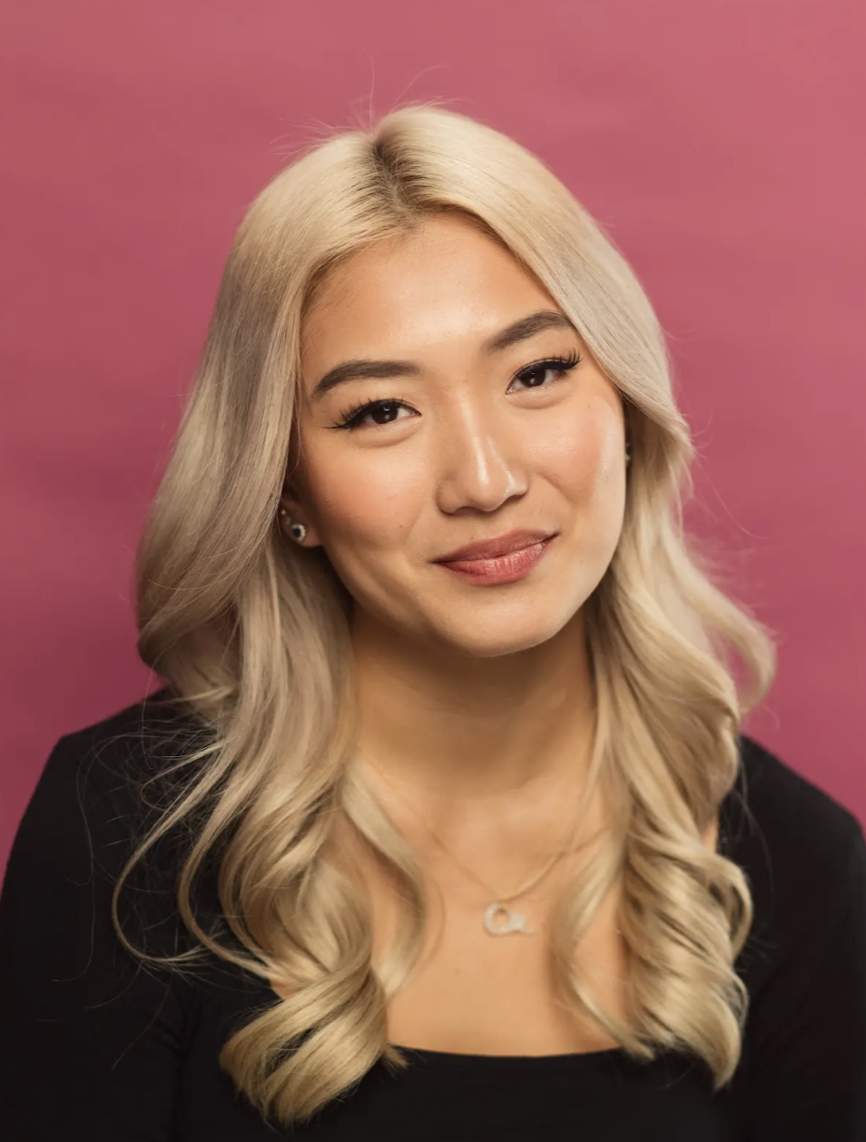 A woman with long, wavy platinum blonde hair, wearing a black top and jewelry, smiling gently against a pink background.