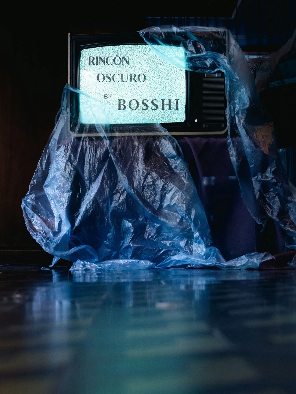 An old television displaying static with text 'RINCÓN OSCURO BY BOSSH' on the screen. The TV is partially wrapped in a plastic sheet, resting on a dark surface, in a dimly lit room.
