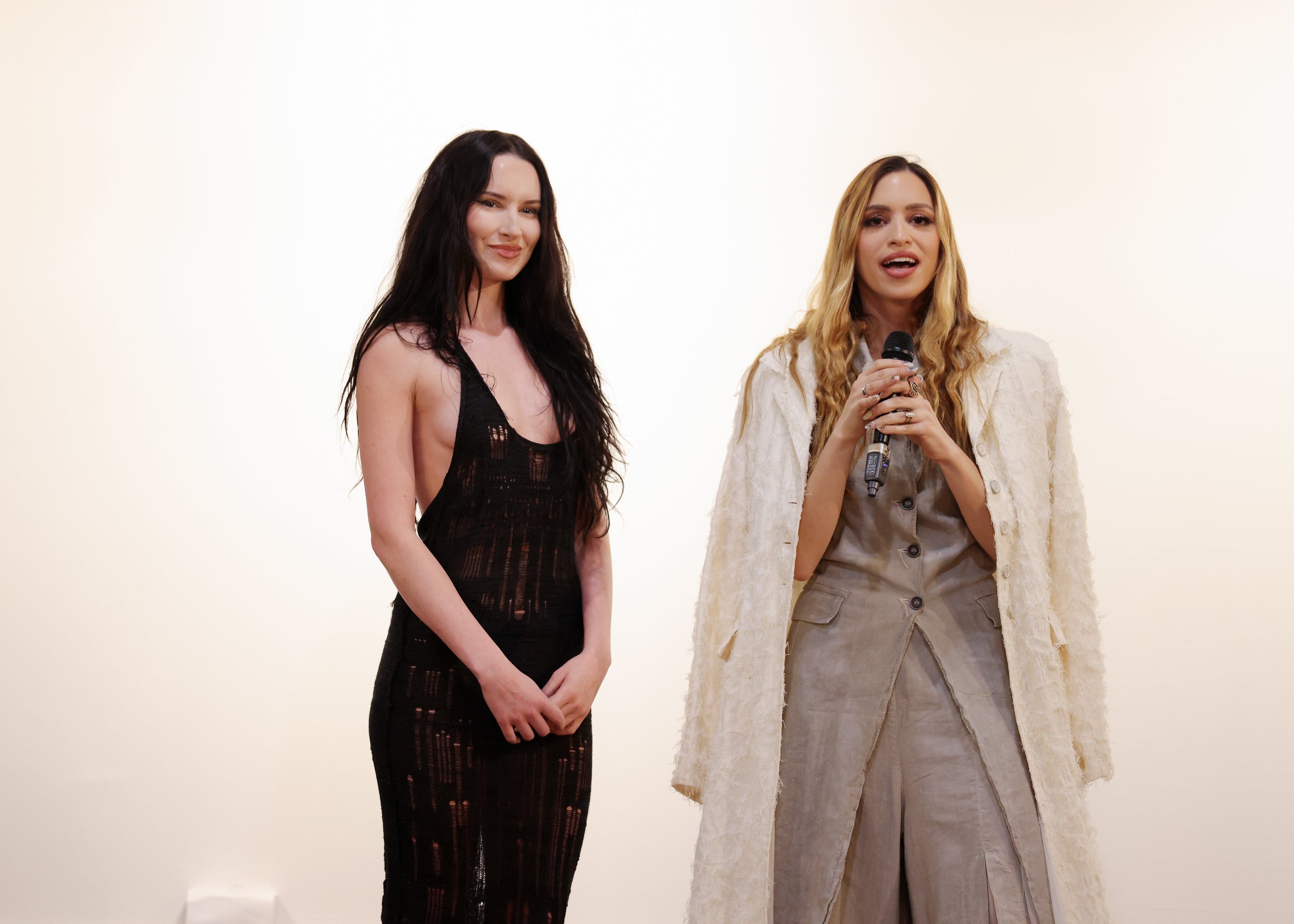 Two women stand against a plain white background; the woman on the left has long dark hair and is wearing a black, sleeveless, fitted dress with cutouts, smiling softly. The woman on the right has long, blonde hair and is holding a microphone, speaking or singing, wearing a light-colored outfit with a textured, off-white coat and beige pants.