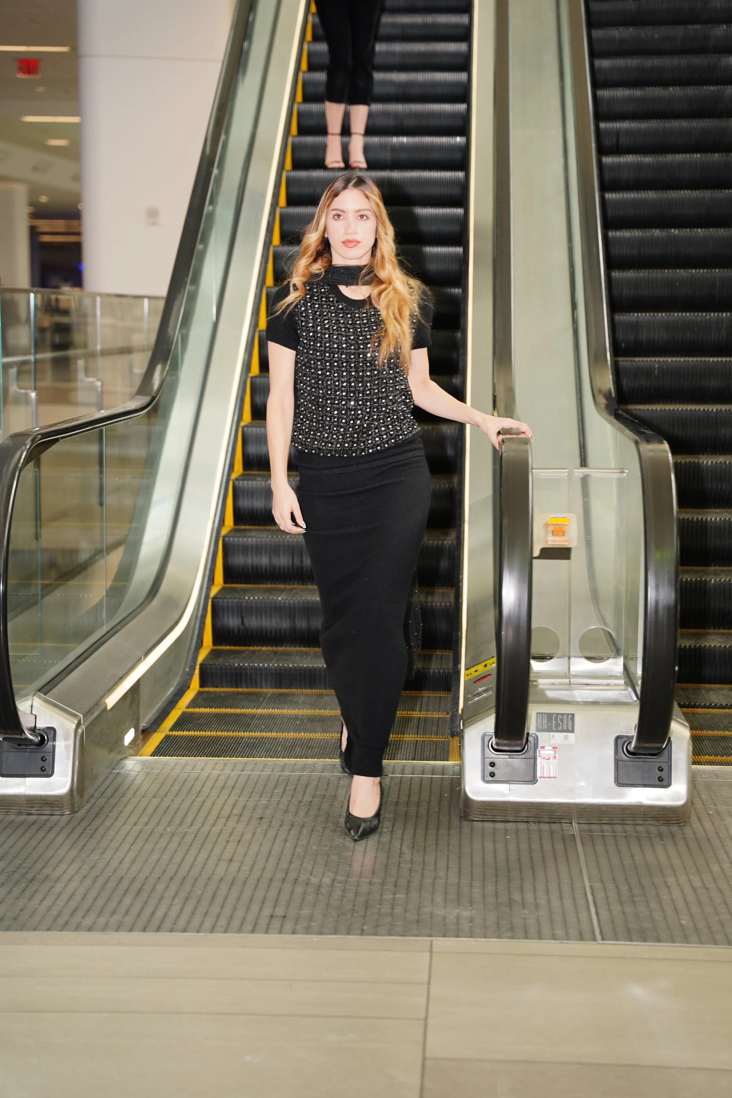 A woman standing on an escalator in a shopping mall, wearing a black dress with a sparkly top and black high heels, holding the handrail with her right hand.