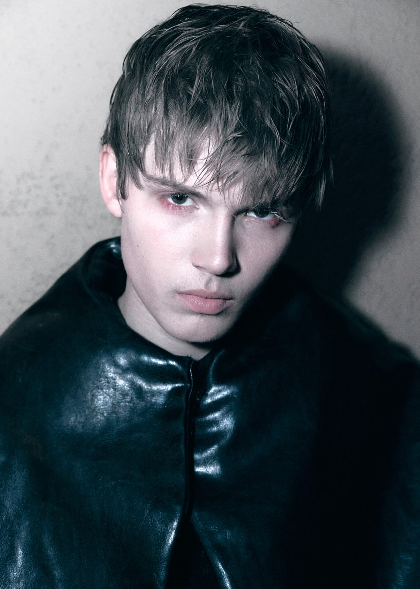 A close-up portrait of a young man with wet hair, wearing a black leather jacket, looking directly at the camera with an intense expression.