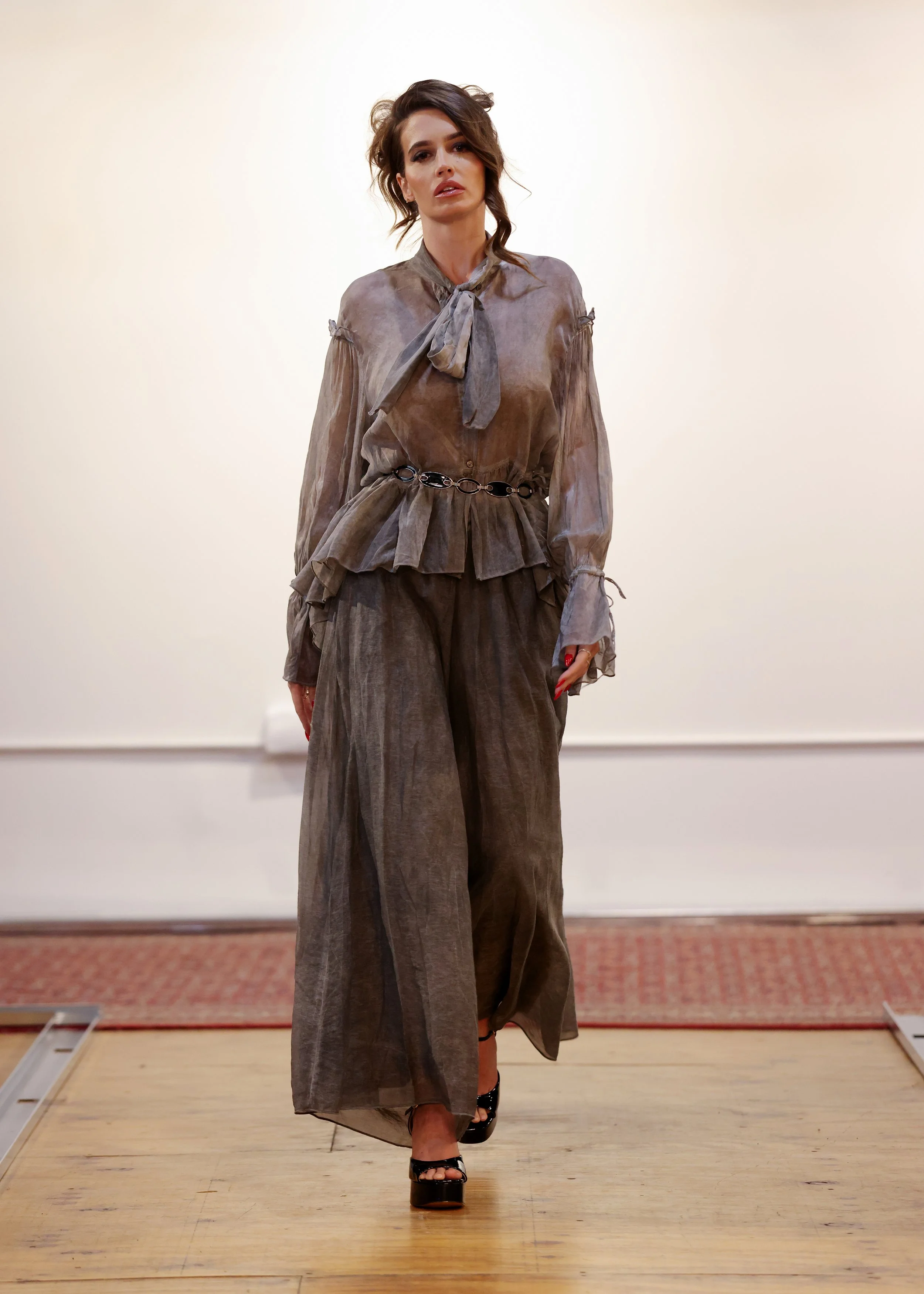 A woman walking on a runway wearing a brown, sheer, long-sleeved blouse with ruffled details and a tie at the collar, paired with wide-legged trousers with a chain belt, and black high-heeled sandals.