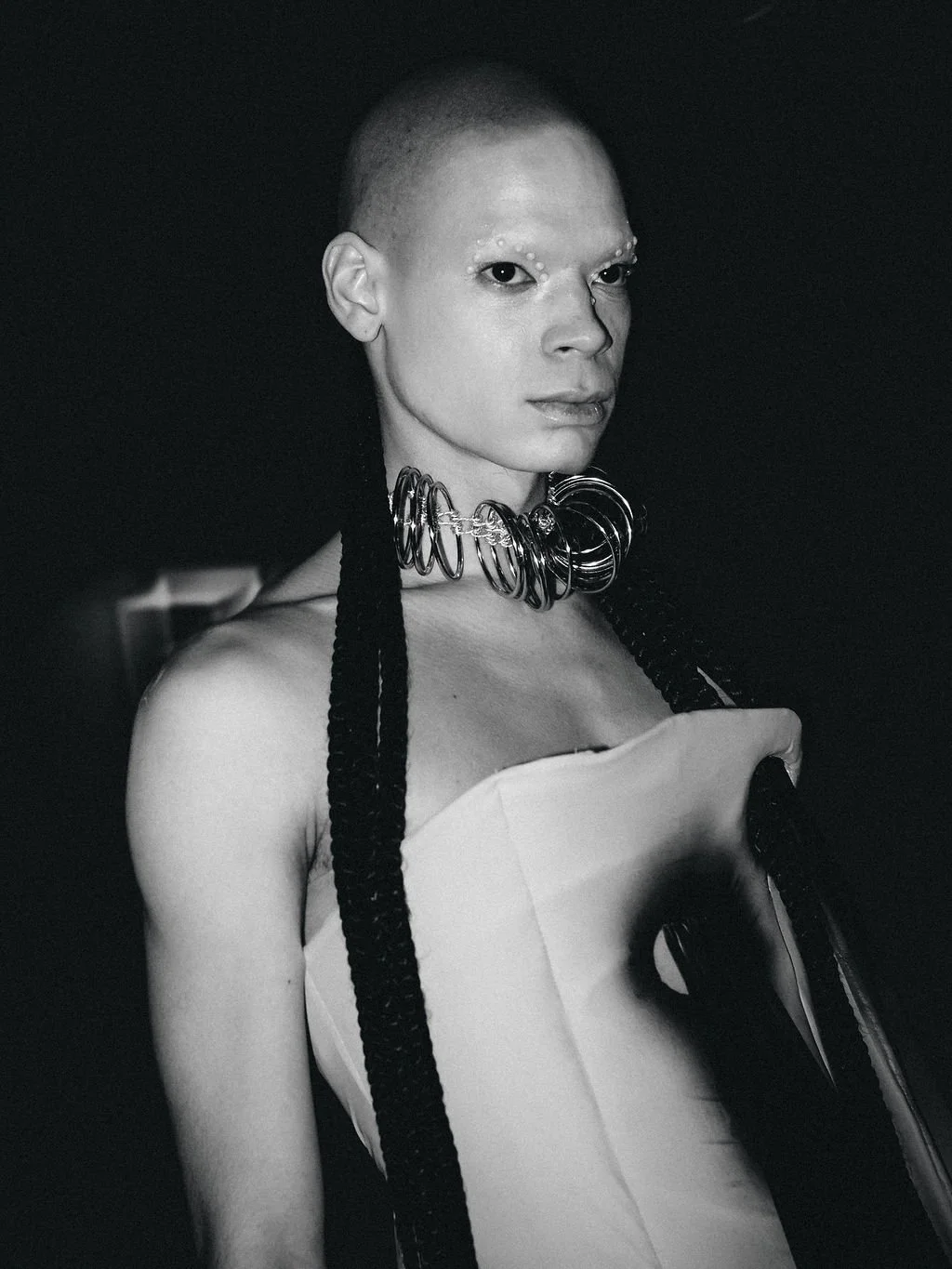 Black and white photo of a person with a shaved head, wearing multiple metallic neck rings, a strapless top, and long braided hair, against a dark background.