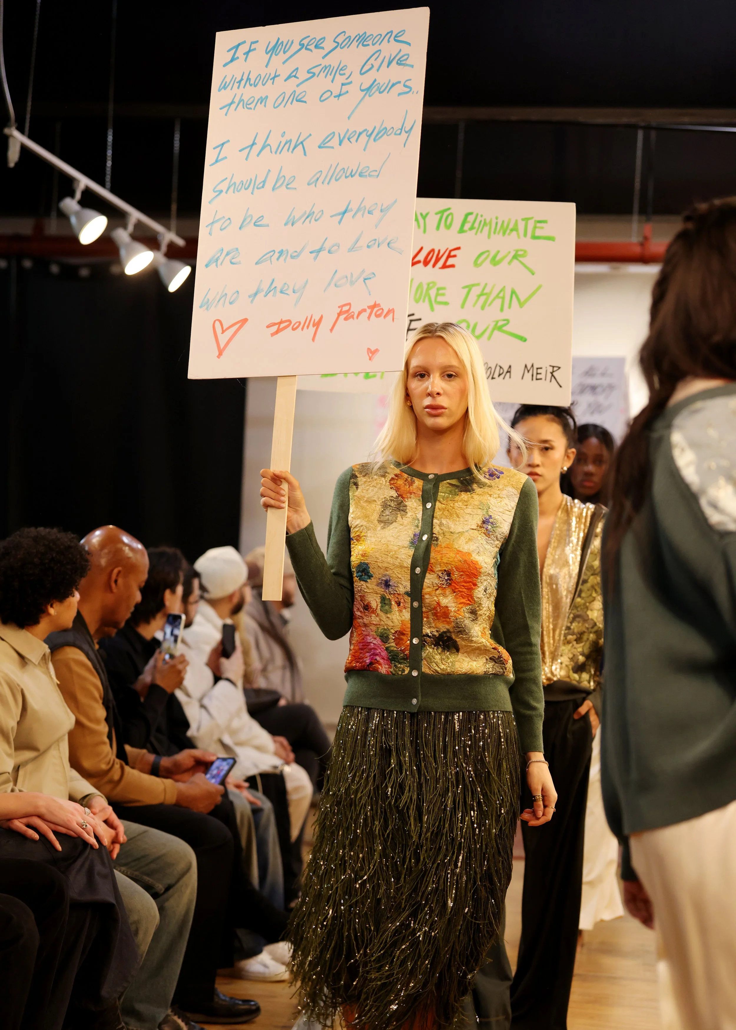 Young woman with blonde hair holding a sign at a protest or rally. The sign reads, 'If you see someone without a smile, give them one of yours... I think everybody should be allowed to be who they are and to love who they love.' The woman is wearing 