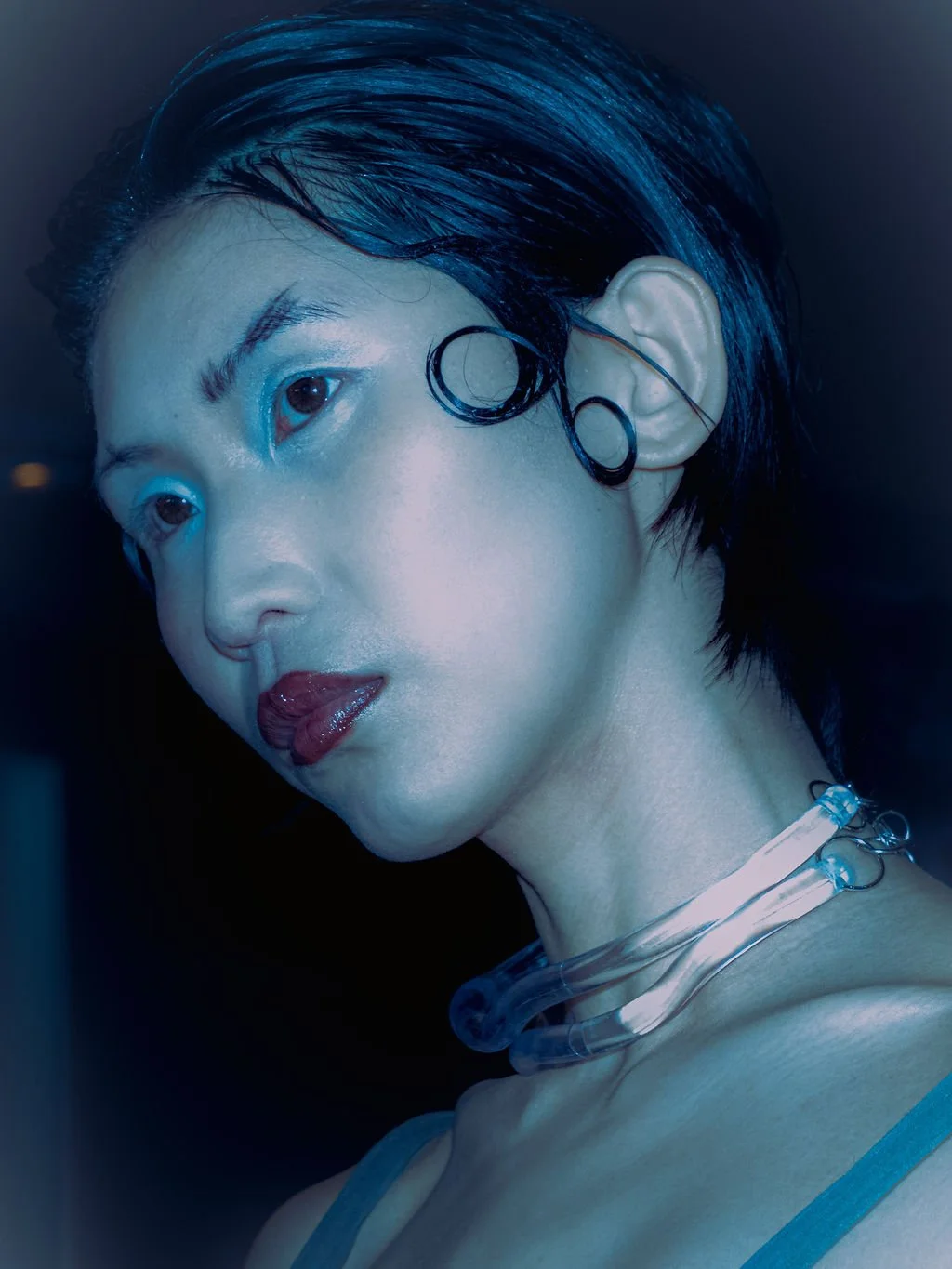 Close-up of a person with short, dark, wet hair, wearing earrings and a necklace, with makeup and lipstick, looking sideways in a dark setting with blue lighting.