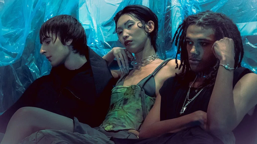 Three young adults sitting closely together against a shiny, blue plastic backdrop, with a moody and stylish appearance.