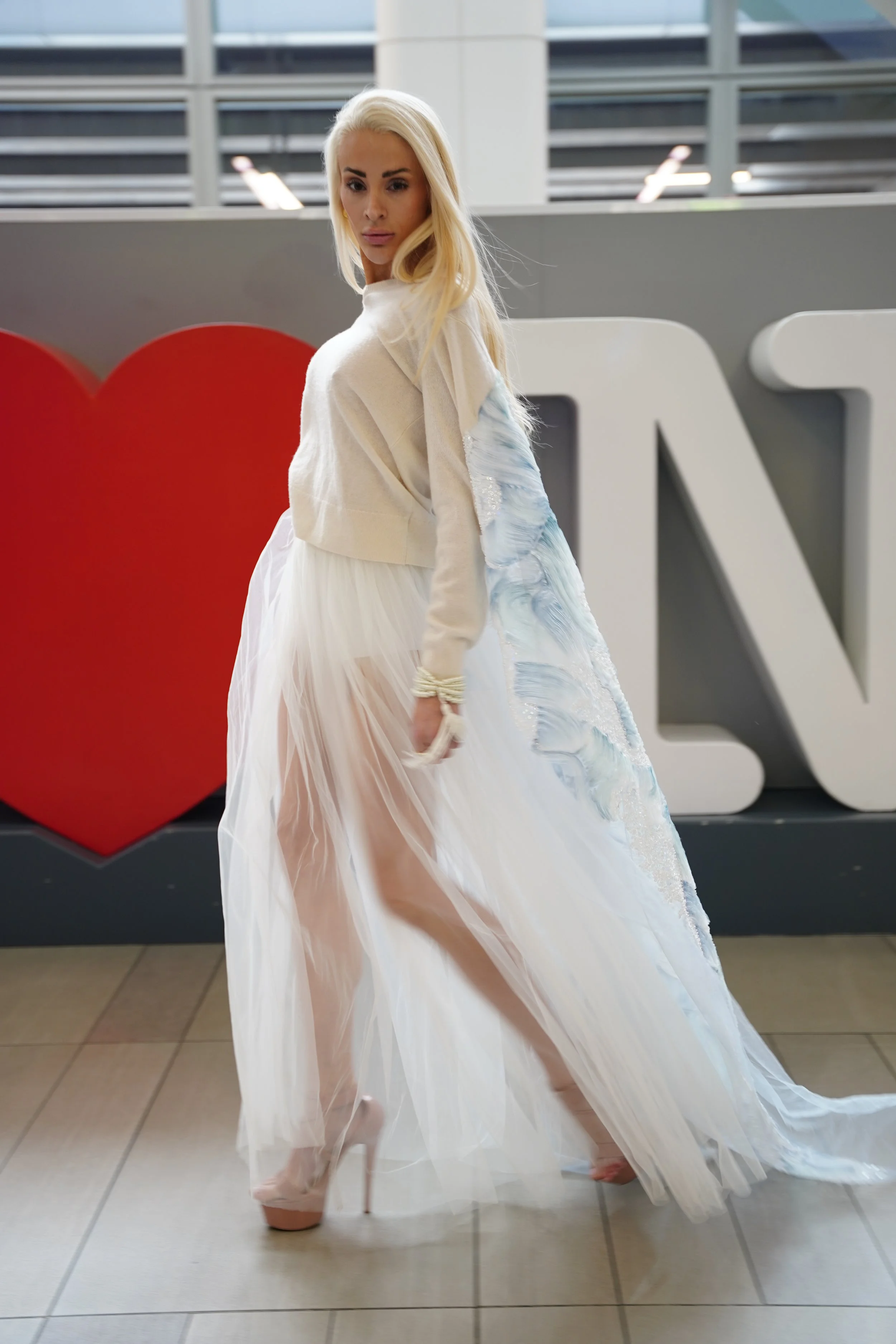 A woman in a white dress standing in an indoor space with large windows, posing in front of a sign with a red heart and white letters.