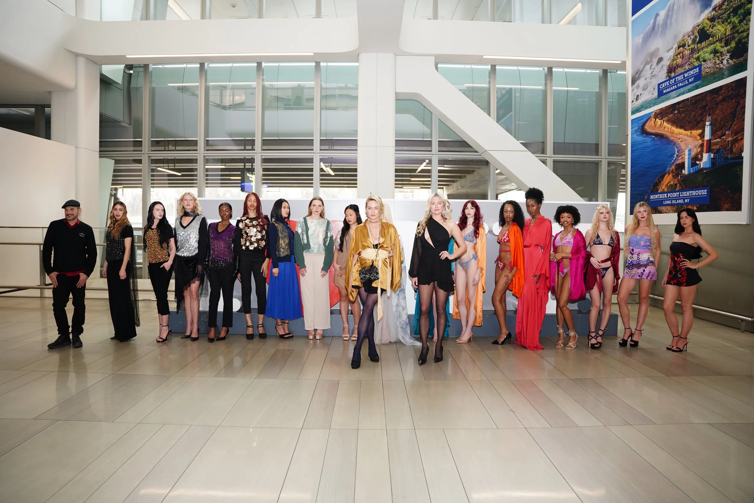 Group of 16 models in fashionable outfits posing in an atrium with modern architecture and large posters of scenic landscapes.