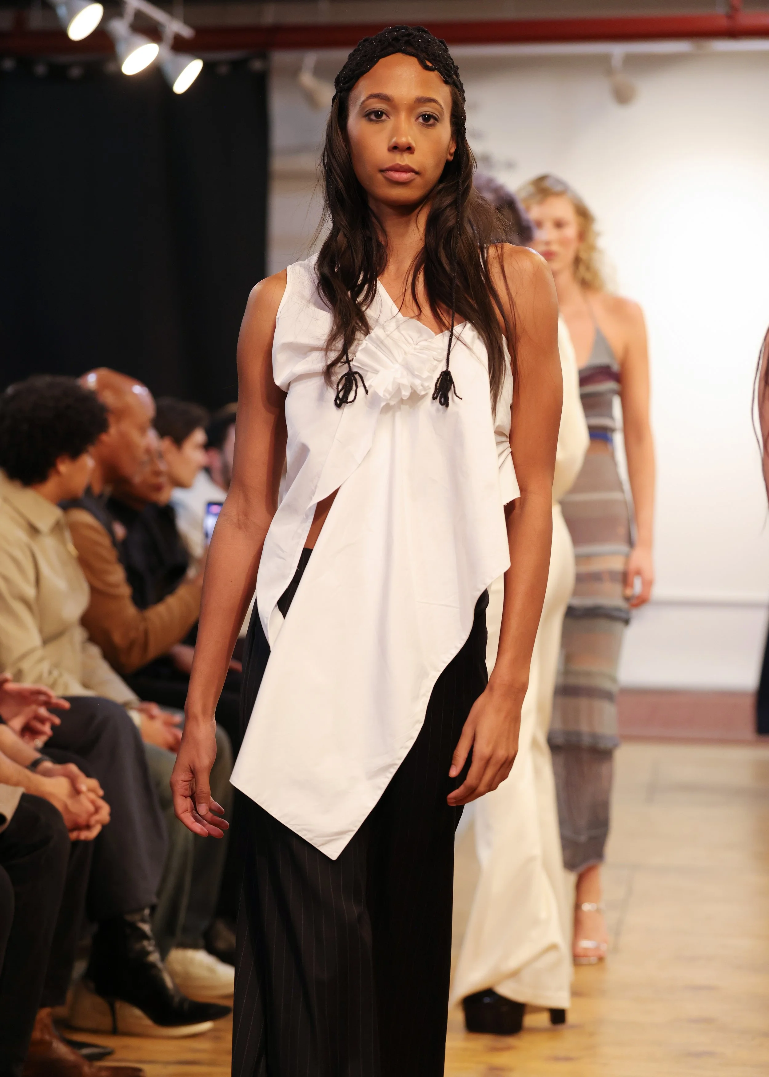 A female model walks down a runway during a fashion show, wearing black pinstripe pants and a white asymmetrical top with black detailing.