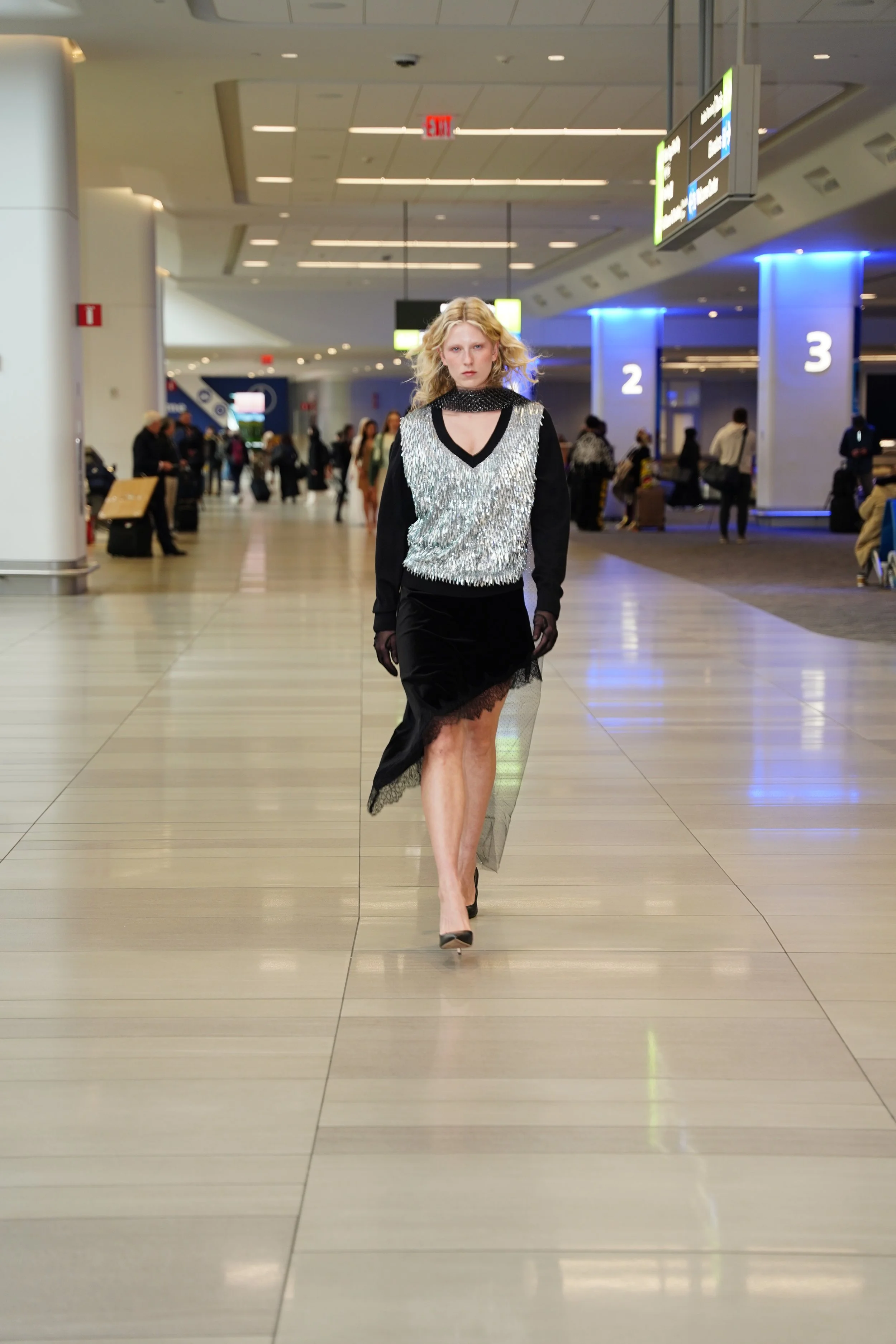 A woman with wavy blonde hair walks in an airport terminal. She is dressed in a stylish, sparkling silver and black dress with sheer lace detailing and black heels.