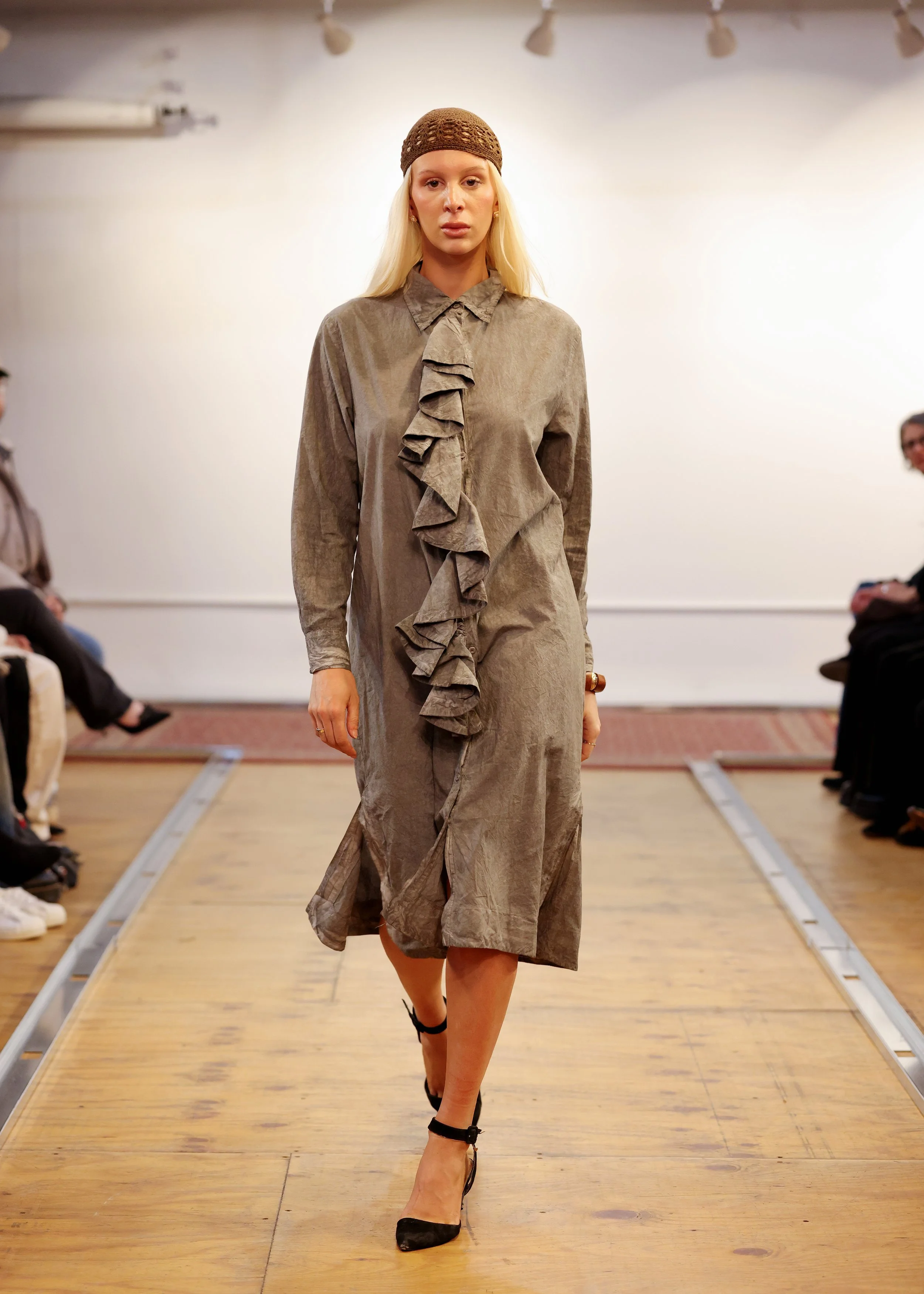 Model walking down runway in taupe-colored dress with ruffle detail, high heels, and a brown knit cap at a fashion show.