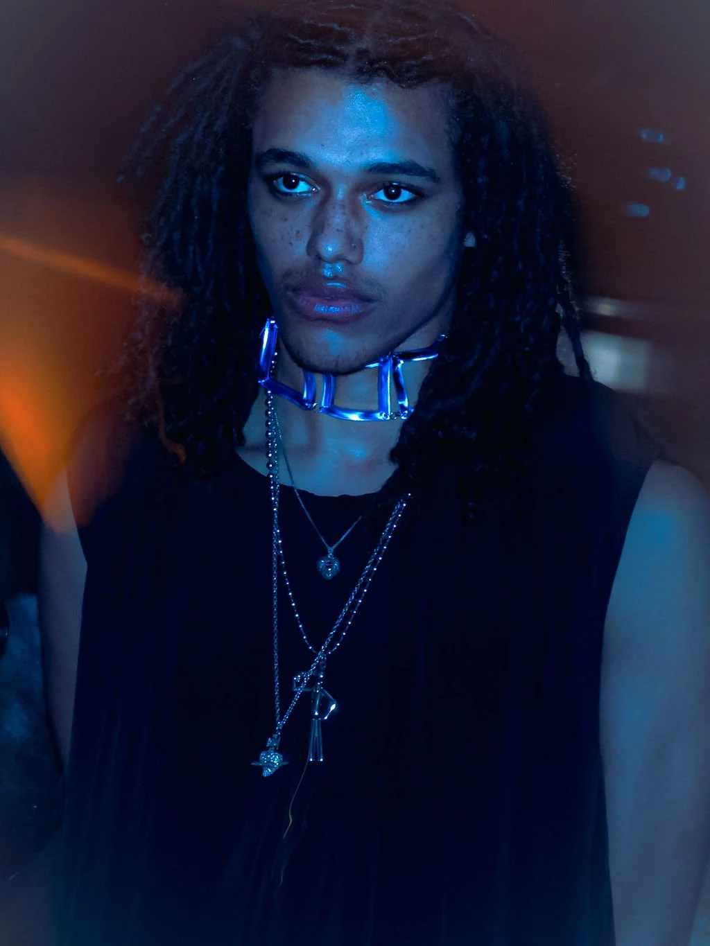 A person with dark, curly hair and multiple necklaces, wearing a black top and a shiny silver choker, standing in a dimly lit environment with blue lighting.