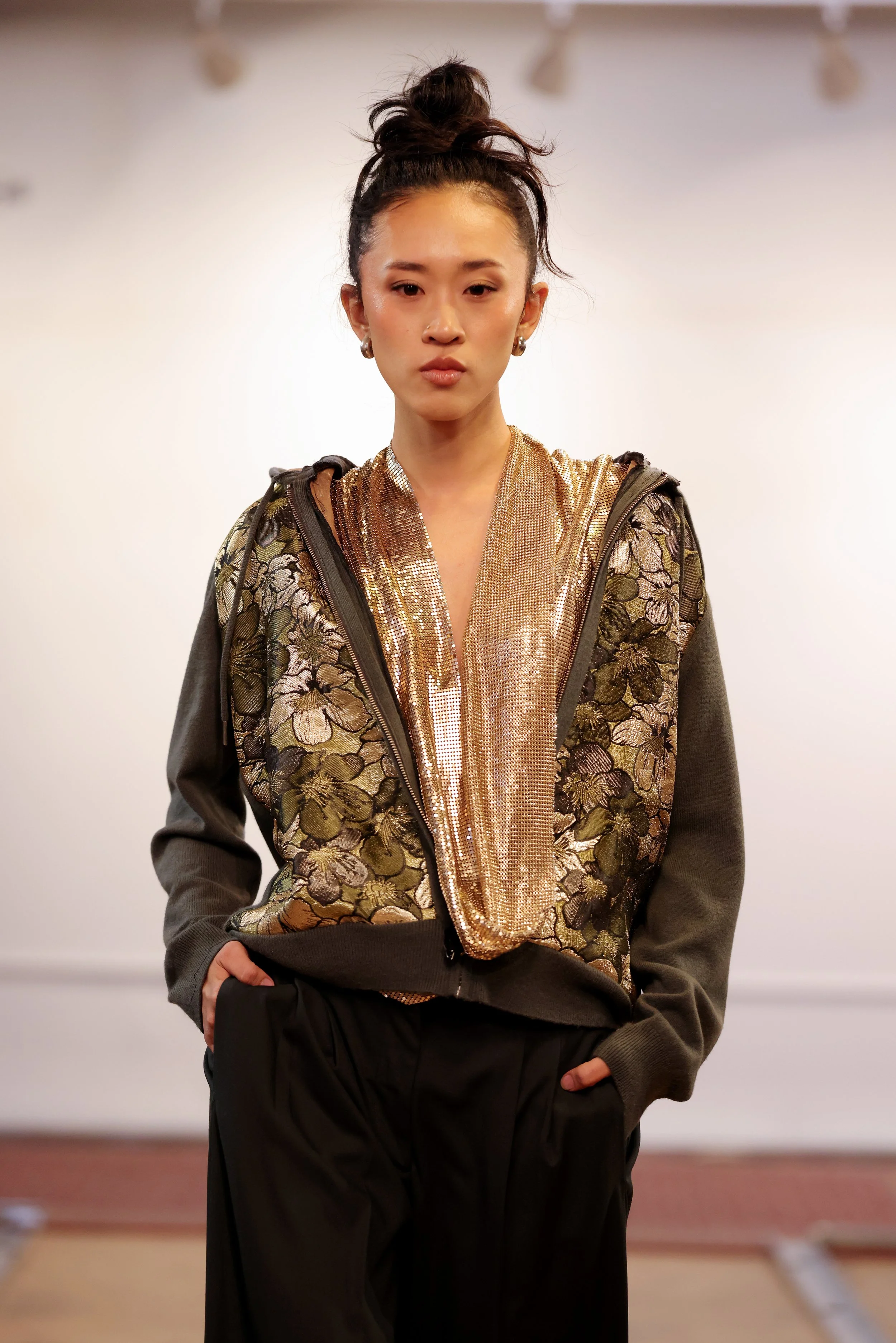 A young woman with dark hair in a messy bun, wearing a sequined top with floral patterns and a dark jacket with zipper details, standing indoors against a plain white wall.