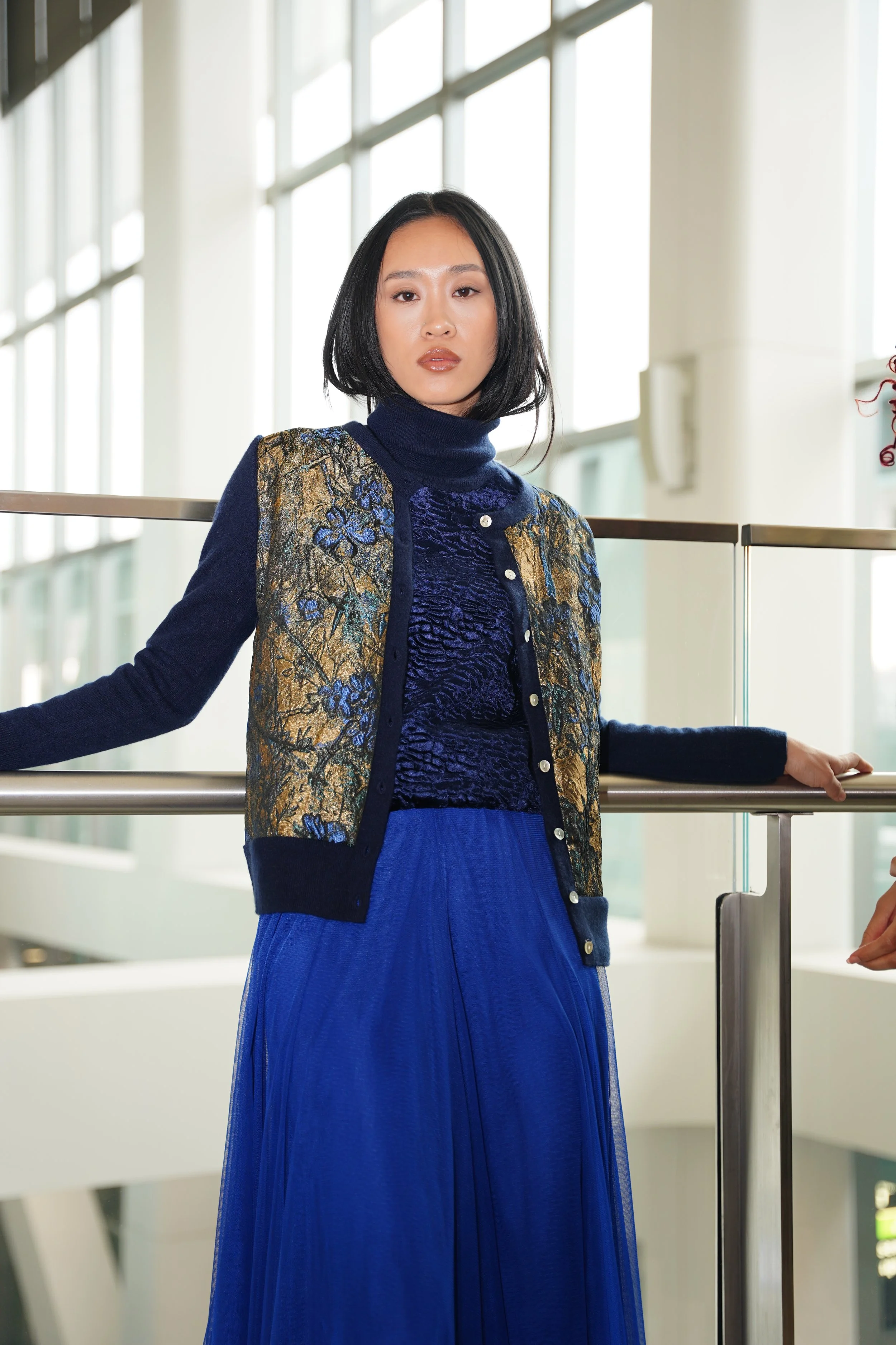 A woman with black shoulder-length hair wearing a blue turtleneck, a patterned vest, and a long blue skirt standing indoors near a glass railing with large window panels in the background.