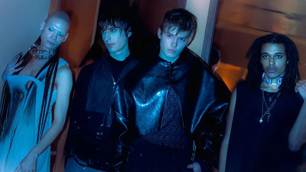 Group of four diverse young adults with fashion-forward outfits, posing in a dimly lit indoor space with a modern design.