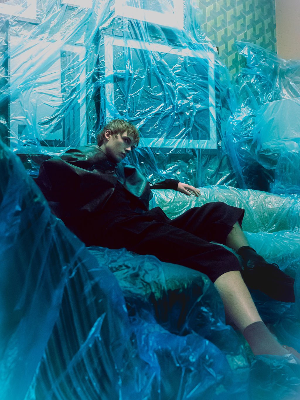 Person sitting on a couch covered with blue plastic, surrounded by plastic sheets and a computer monitor in the background.