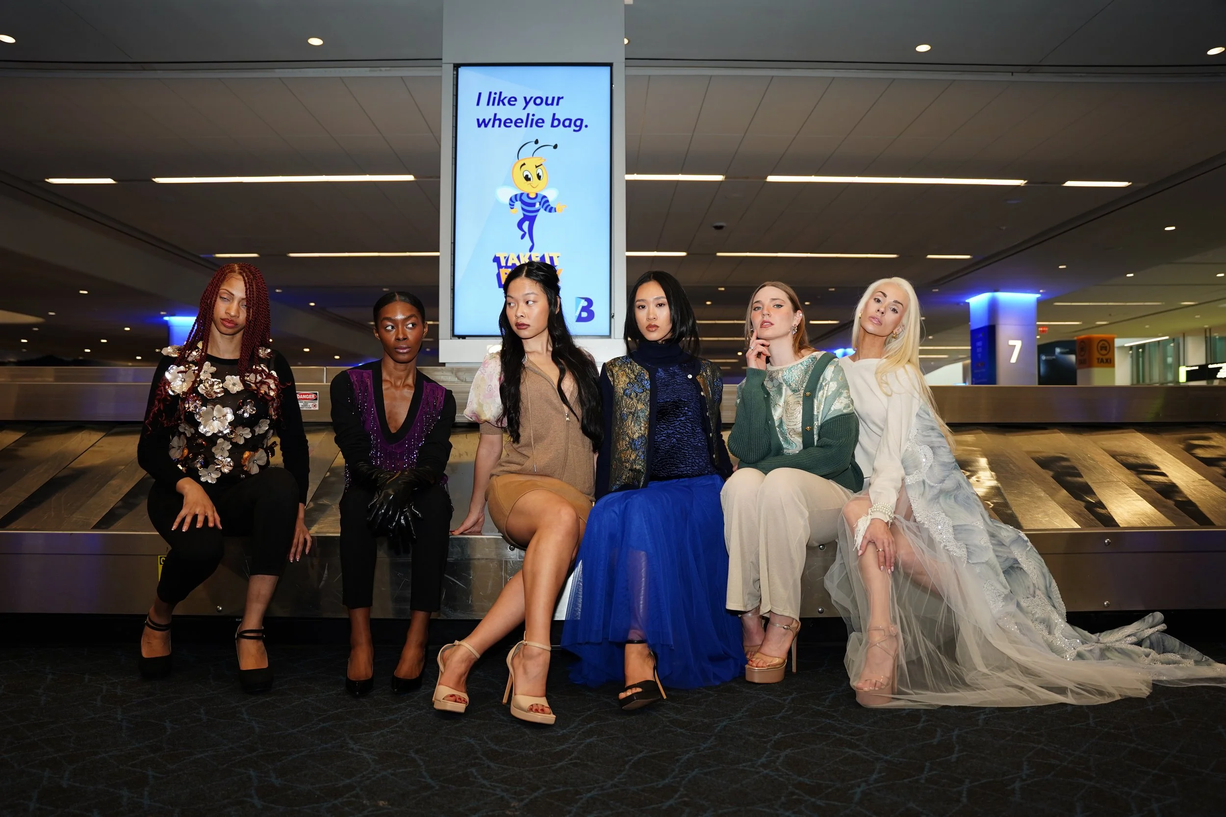 A group of six women sitting on a luggage carousel at an airport, posing for a photo. They are dressed in various fashionable outfits, with a digital display screen behind them showing a cartoon bee and text that says, 'I like your wheelie bag. Take 