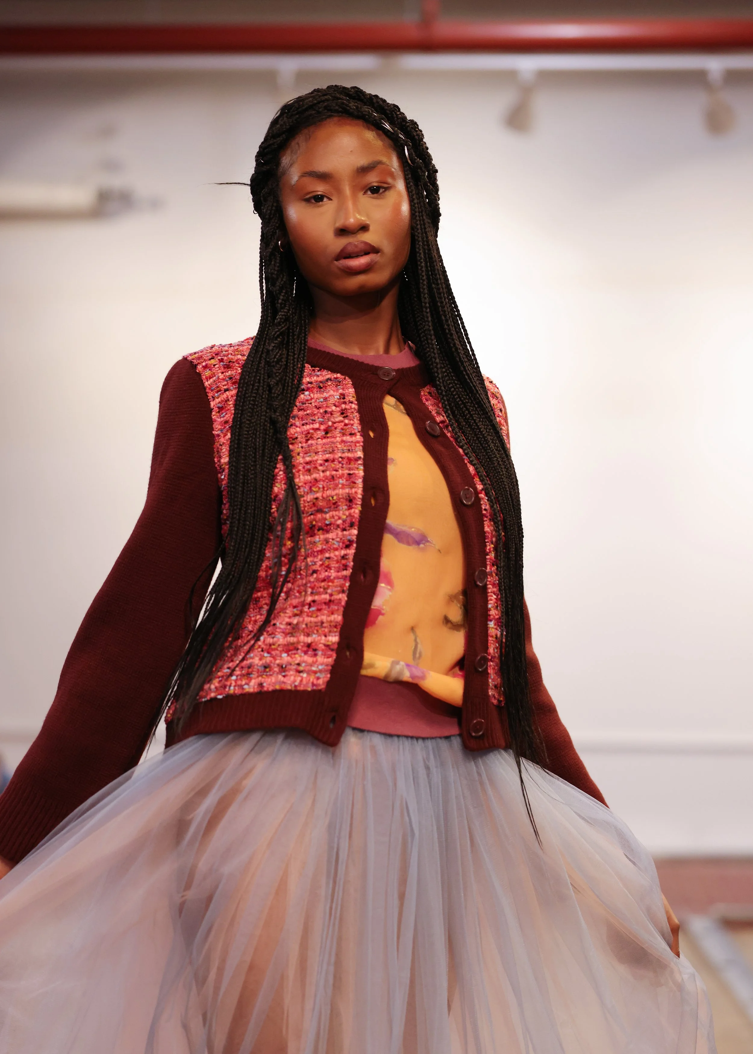 A woman with long braided hair wearing a multicolored jacket and a tulle skirt, standing in an indoor setting with a plain background.
