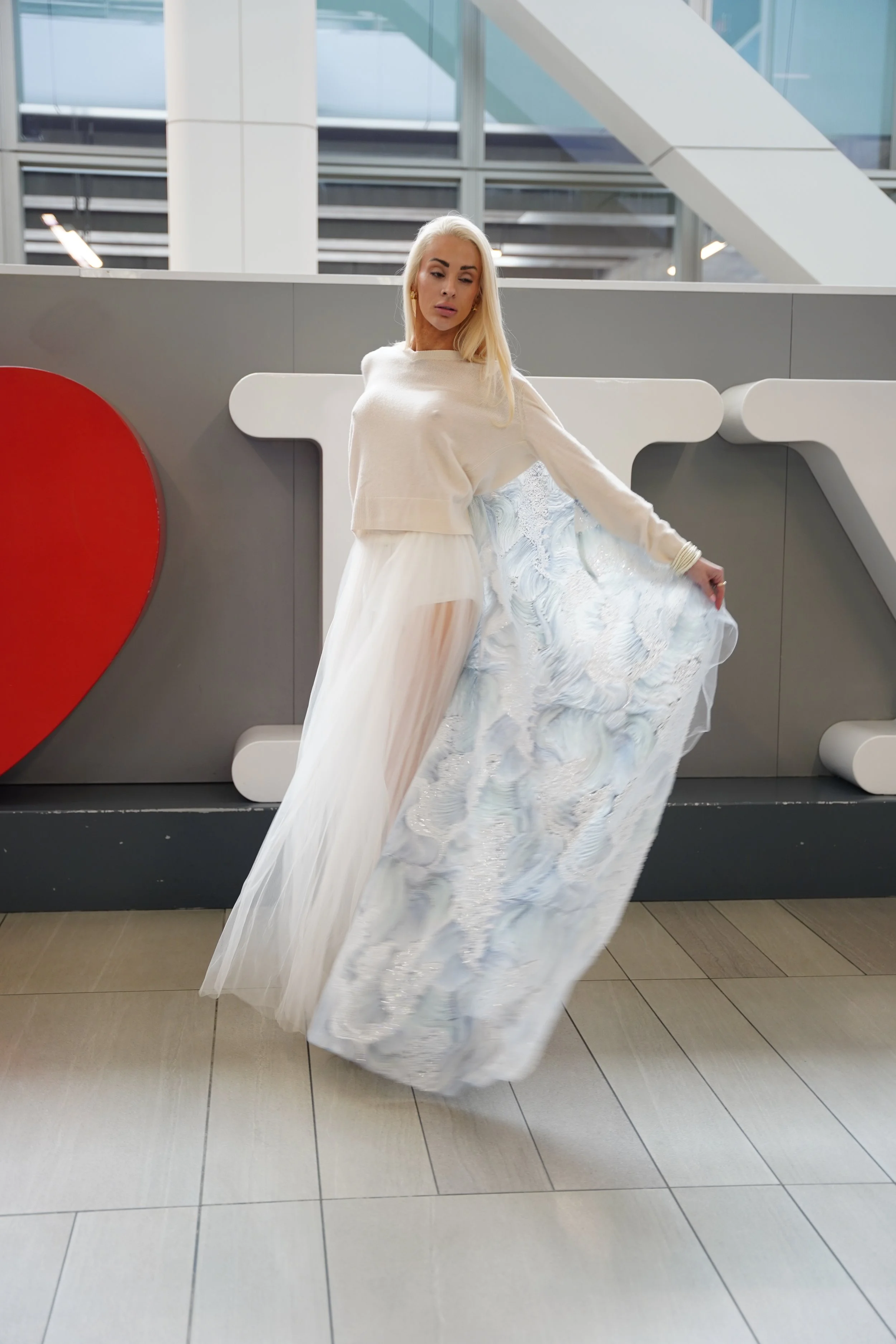 A woman with long blonde hair wearing a white dress with a sheer, flowing skirt, is standing indoors with modern architecture, glass walls, and a large red heart shape partially visible behind her.