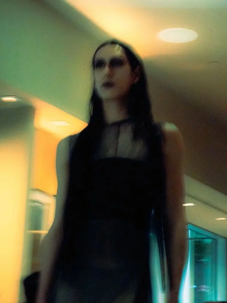 A woman with pale skin, dark makeup, and long dark hair standing in a dimly lit indoor setting.