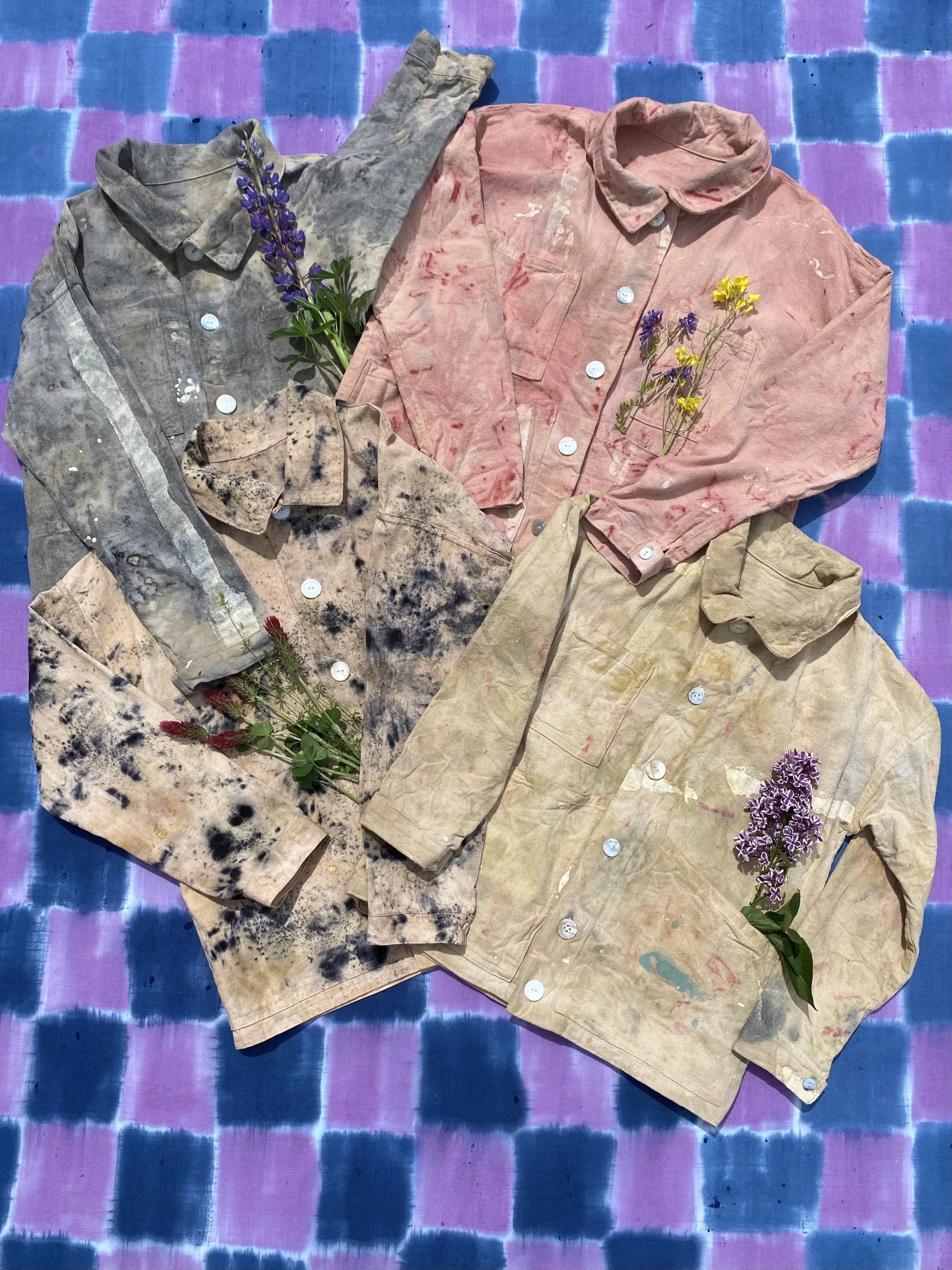 Several worn and stained dress shirts in various colors laid out on a pink and blue plaid fabric surface with sprigs of purple and yellow flowers placed on them.