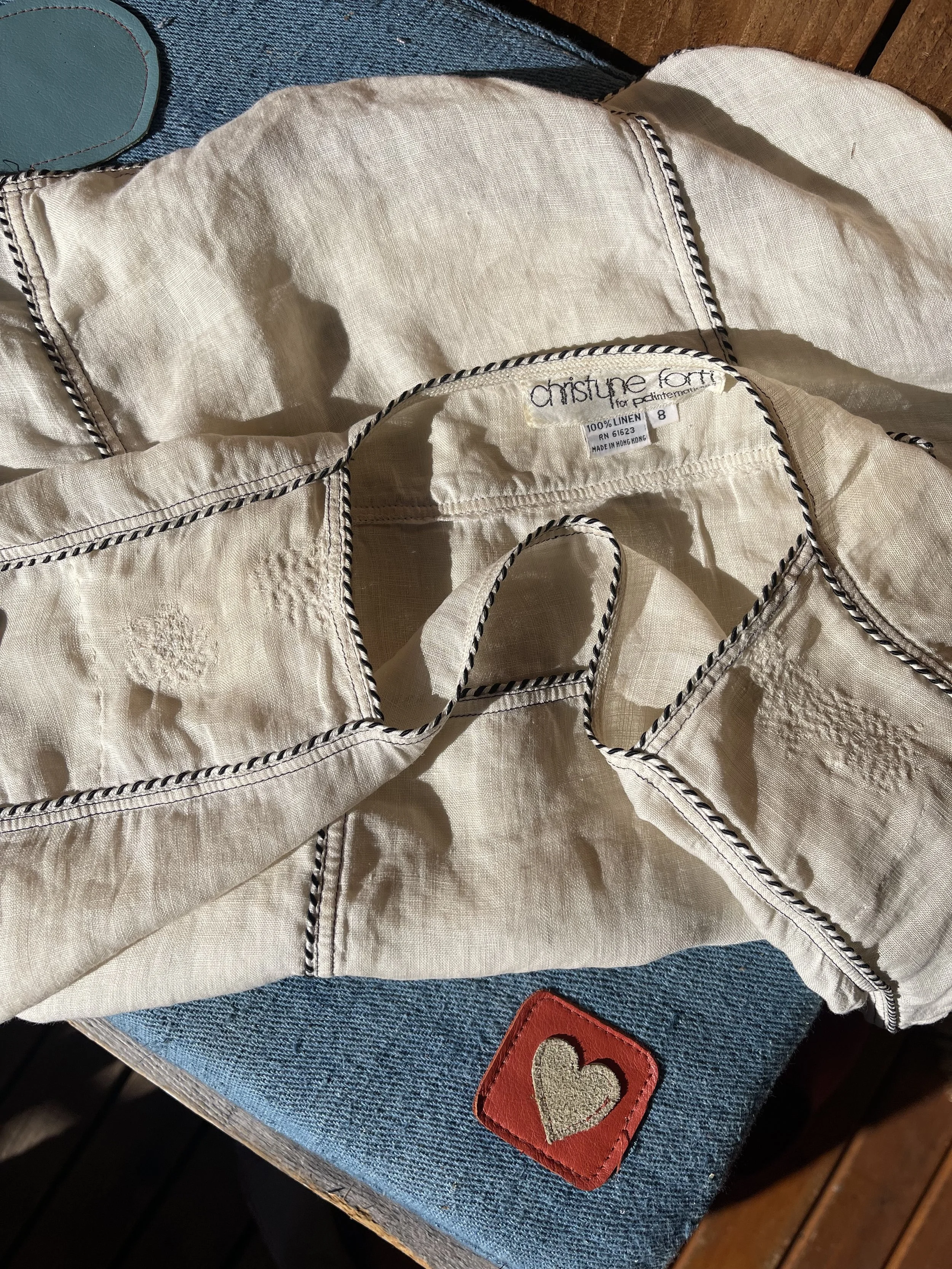 A cream-colored linen shirt with black and white twisted piping, lying on a blue textured fabric surface. The shirt has a tag that reads 'Christine Fort for peainting', made of 100% linen, size 8, made in Hong Kong. There is a red leather patch with 