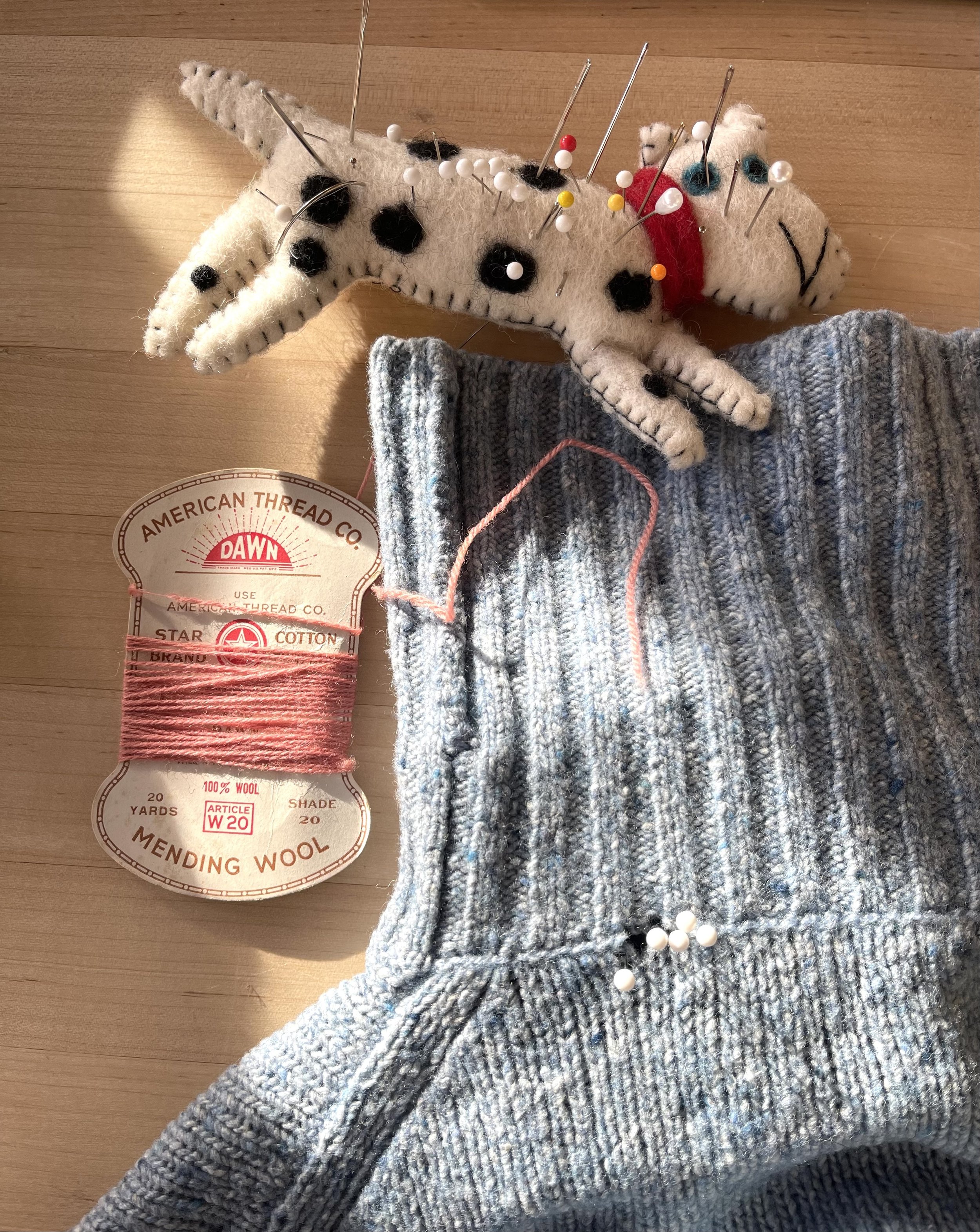 A small plush animal resembling a dog with black spots, closed eyes, and a red collar, lying on a flat surface with numerous sewing pins inserted into it. Nearby, there is a spool of pink thread and a piece of blue and gray knitted fabric.
