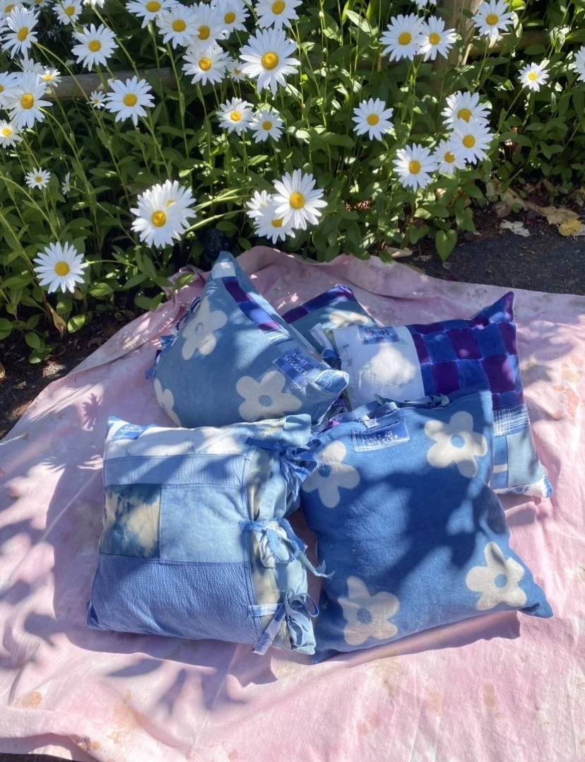 Multiple blue and white tie-dye fabric bundles, pillows, and blankets rest on a pink cloth on the ground, with white daisies with yellow centers in the background.