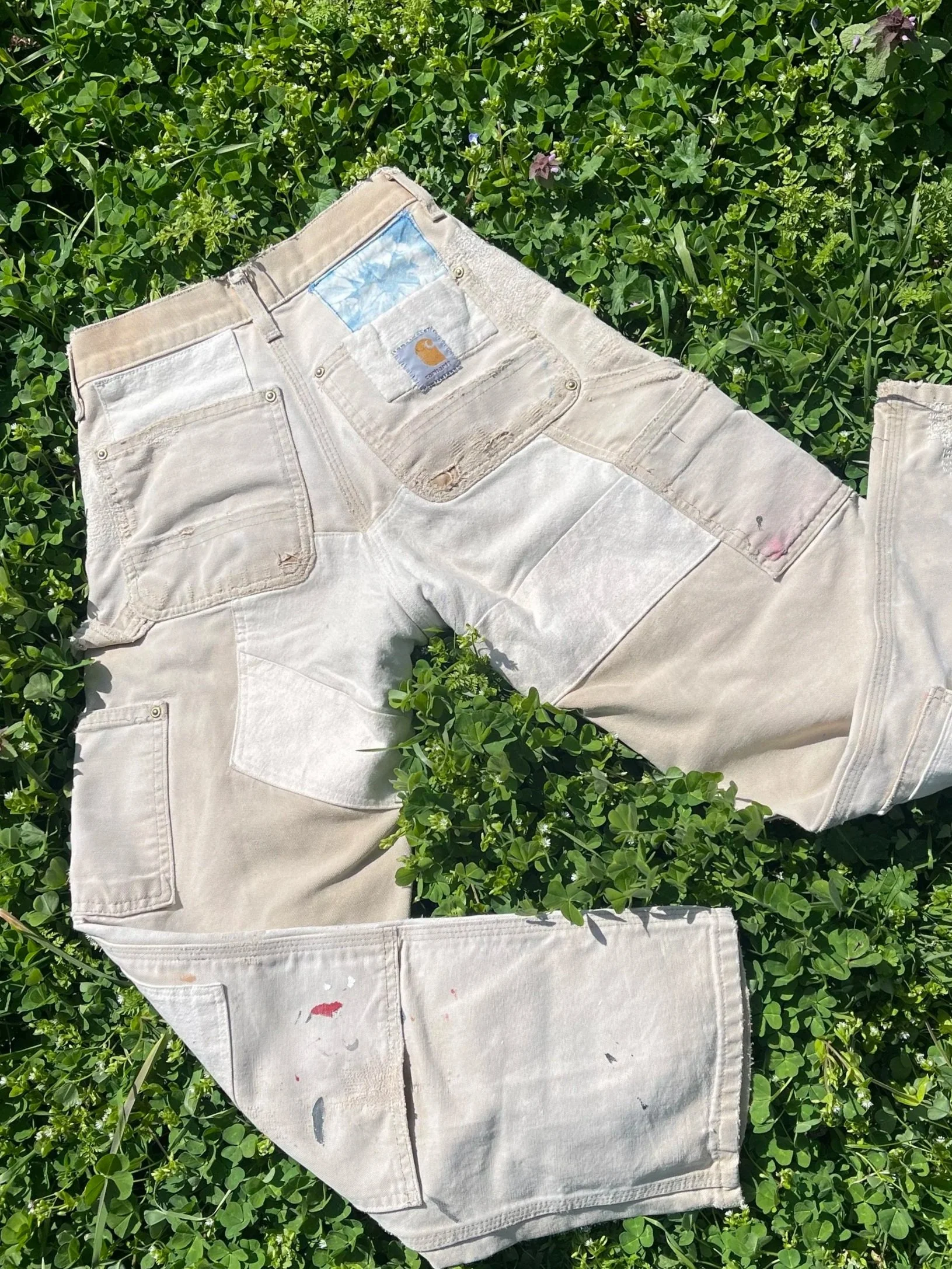A pair of white distressed jeans with paint splatters lying on green grass and clovers.