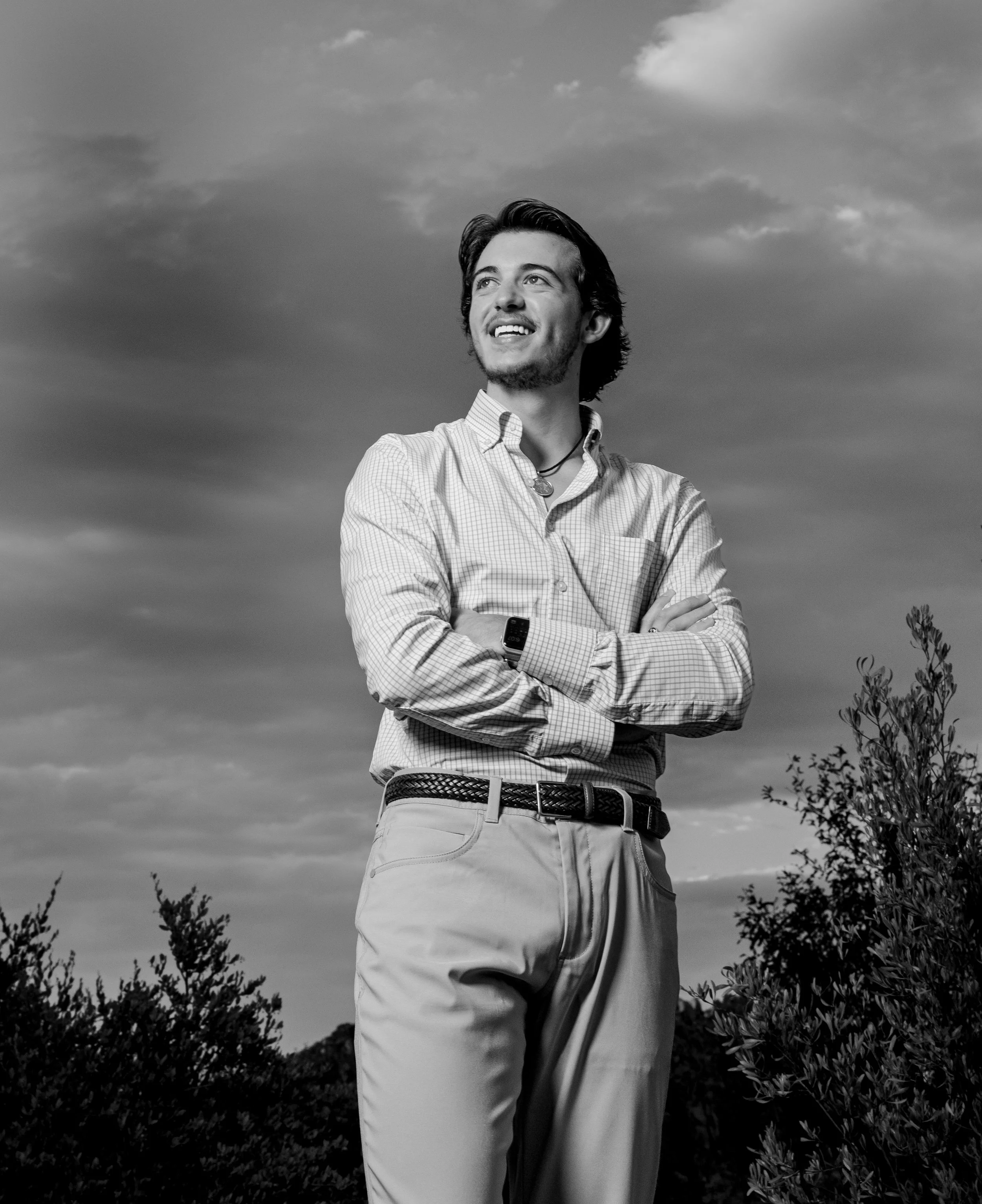 A young man standing outdoors with his arms crossed, smiling and looking off into the distance. He is wearing a button-up shirt, khaki pants, and a smartwatch, with clouds and trees in the background.