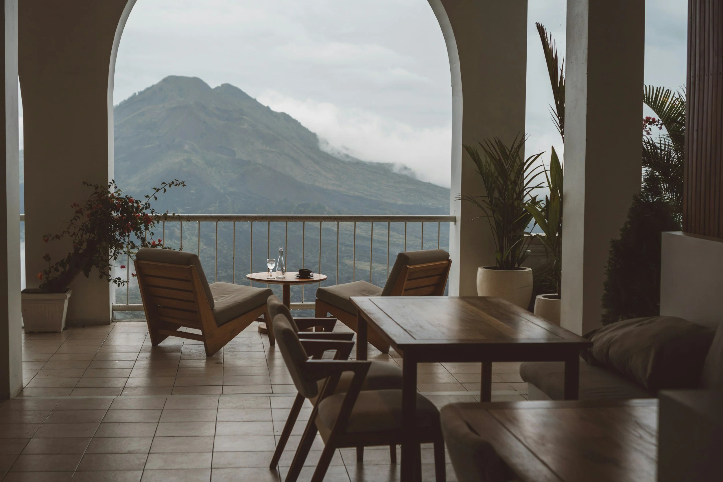 Indoor seating area with wooden furniture and plants, overlooking a mountain through an archway.