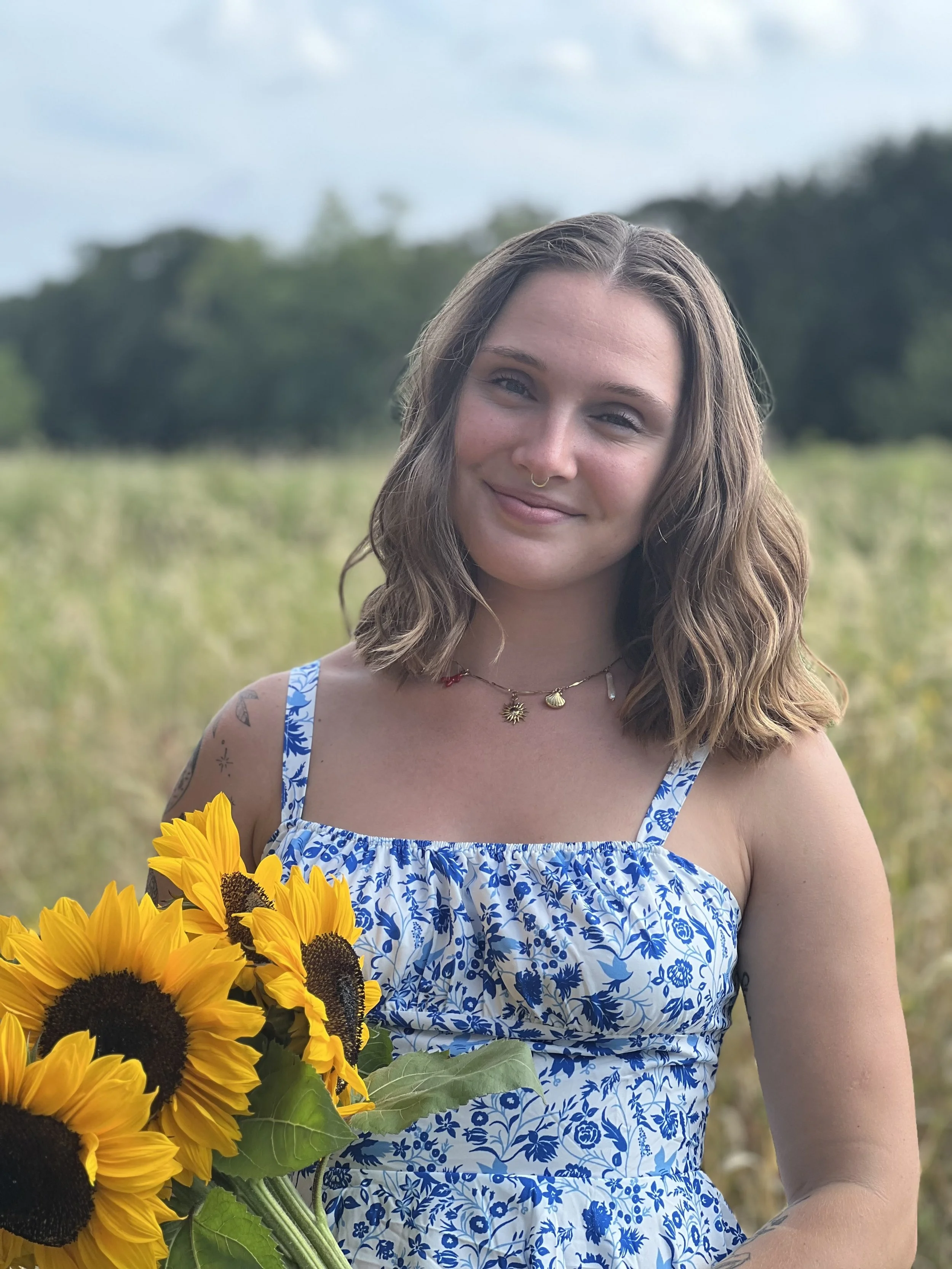 A young woman with shoulder-length wavy hair, wearing a blue and white floral dress, holding a bouquet of sunflowers, standing outdoors in a field with a blurred green background.