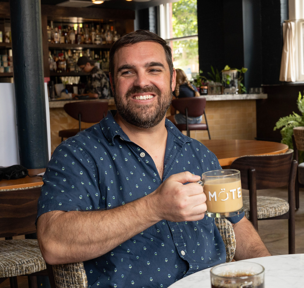 Mote co-founder and head roaster Jonathan Beardsley - a man smiling and holding a mug at a table in a cozy cafe