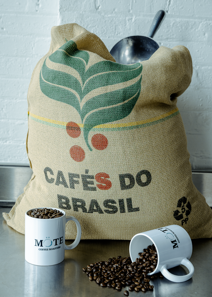 A burlap bag with the logo "Cafés do Brasil" and a green leaf design, filled with coffee beans and a metal scoop sticking out. Two white coffee mugs labeled "Mote Coffee Roasters" are in front, one filled with coffee beans and the other spilled onto the surface.
