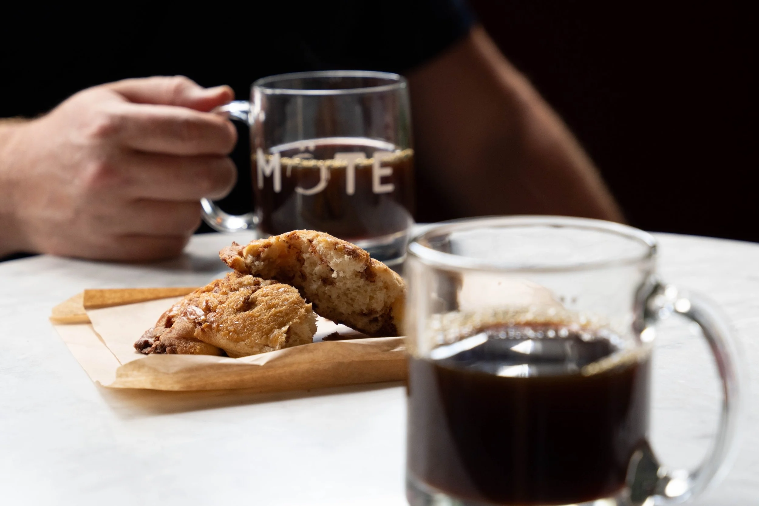 Close-up of a person's hand holding a glass mug of coffee or tea, with cookies and a small glass jar on a white surface in the foreground.