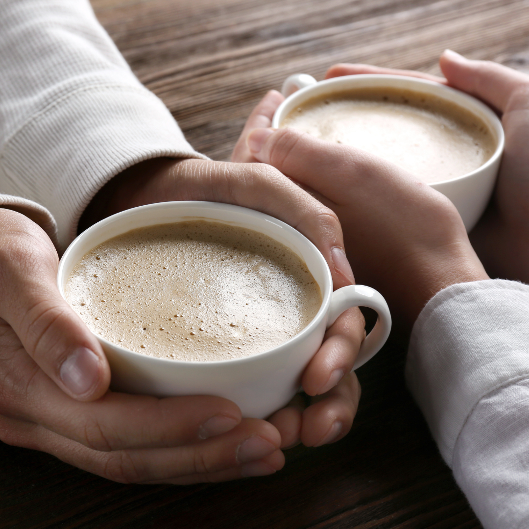 Two people holding white mugs filled with frothy coffee or cappuccino, resting on a wooden table.