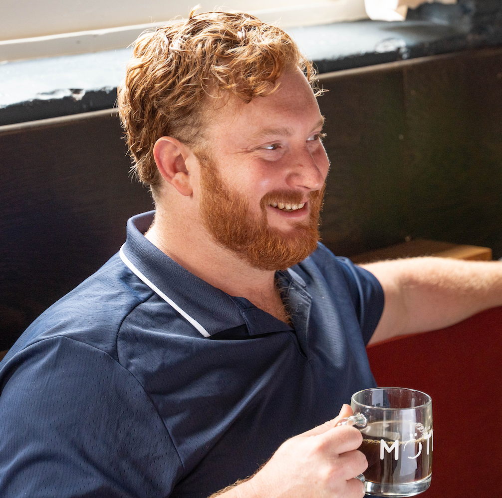 A red-haired man with a beard smiling while holding a clear mug of coffee, sitting indoors near a window.