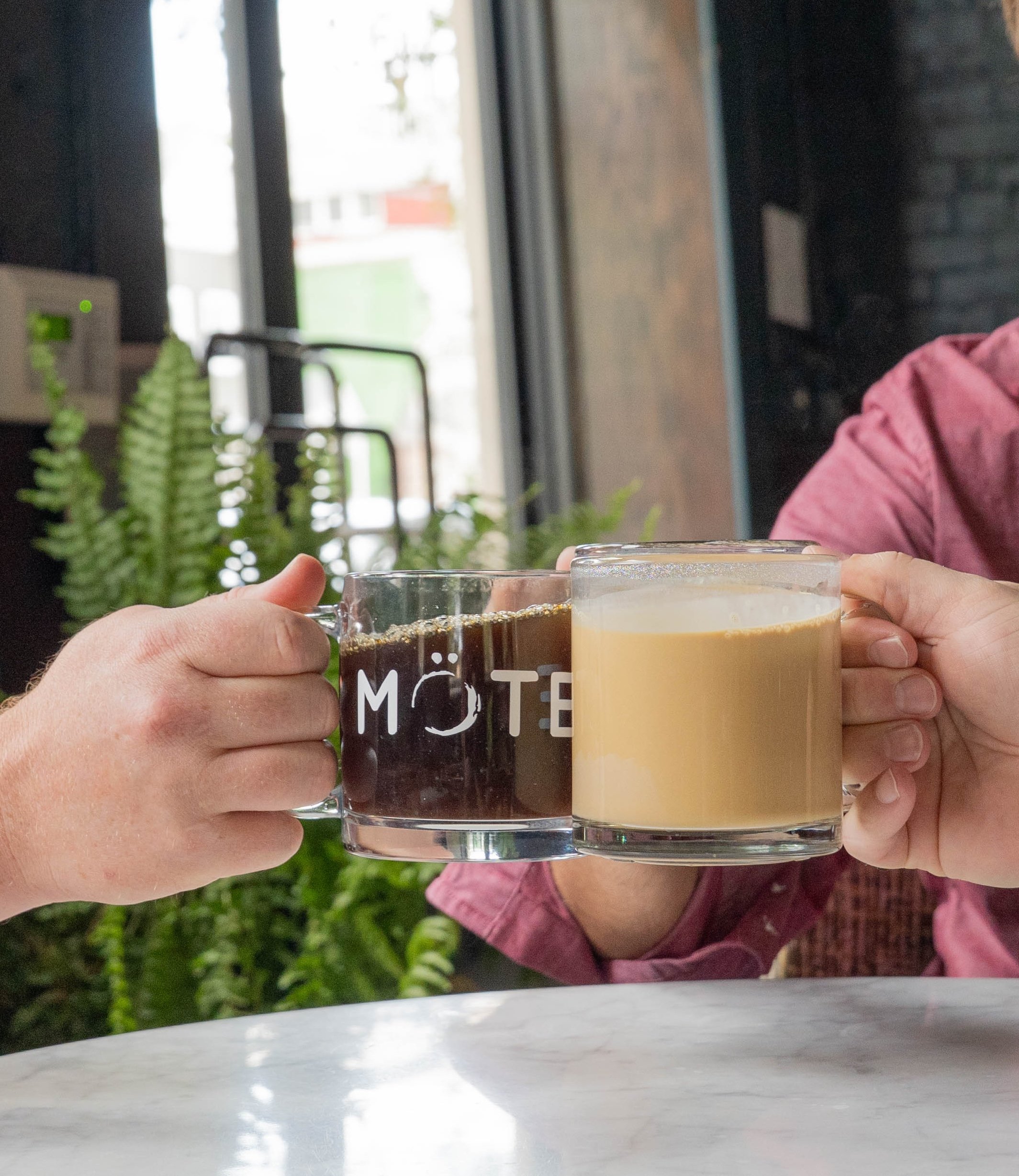 Two hands holding mugs, one black with white text and the other light brown, at a table in front of a window with greenery outside.