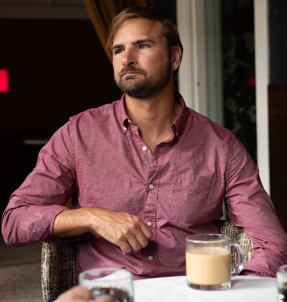 Mote co-founder and CEO Justin Kumor - man with brown hair and a beard sitting at a table with a glass mug of coffee wearing a red button-up shirt at a cozy indoor space