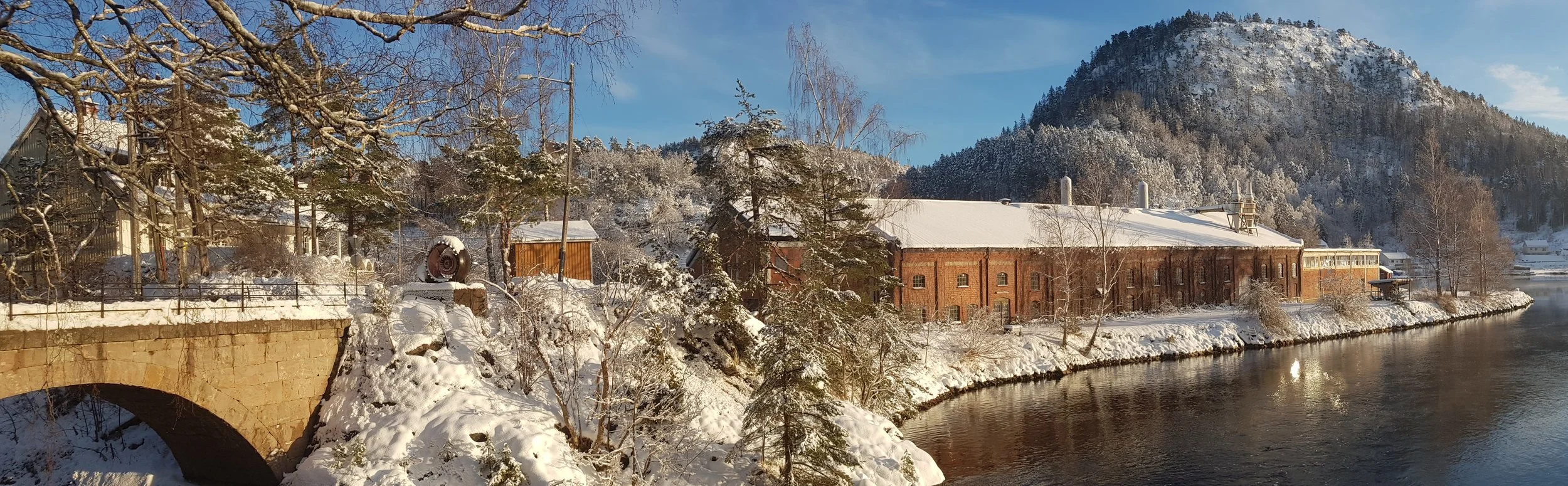 Snow-covered river with brick building and trees on the riverbank, mountains in the background, and a blue sky.