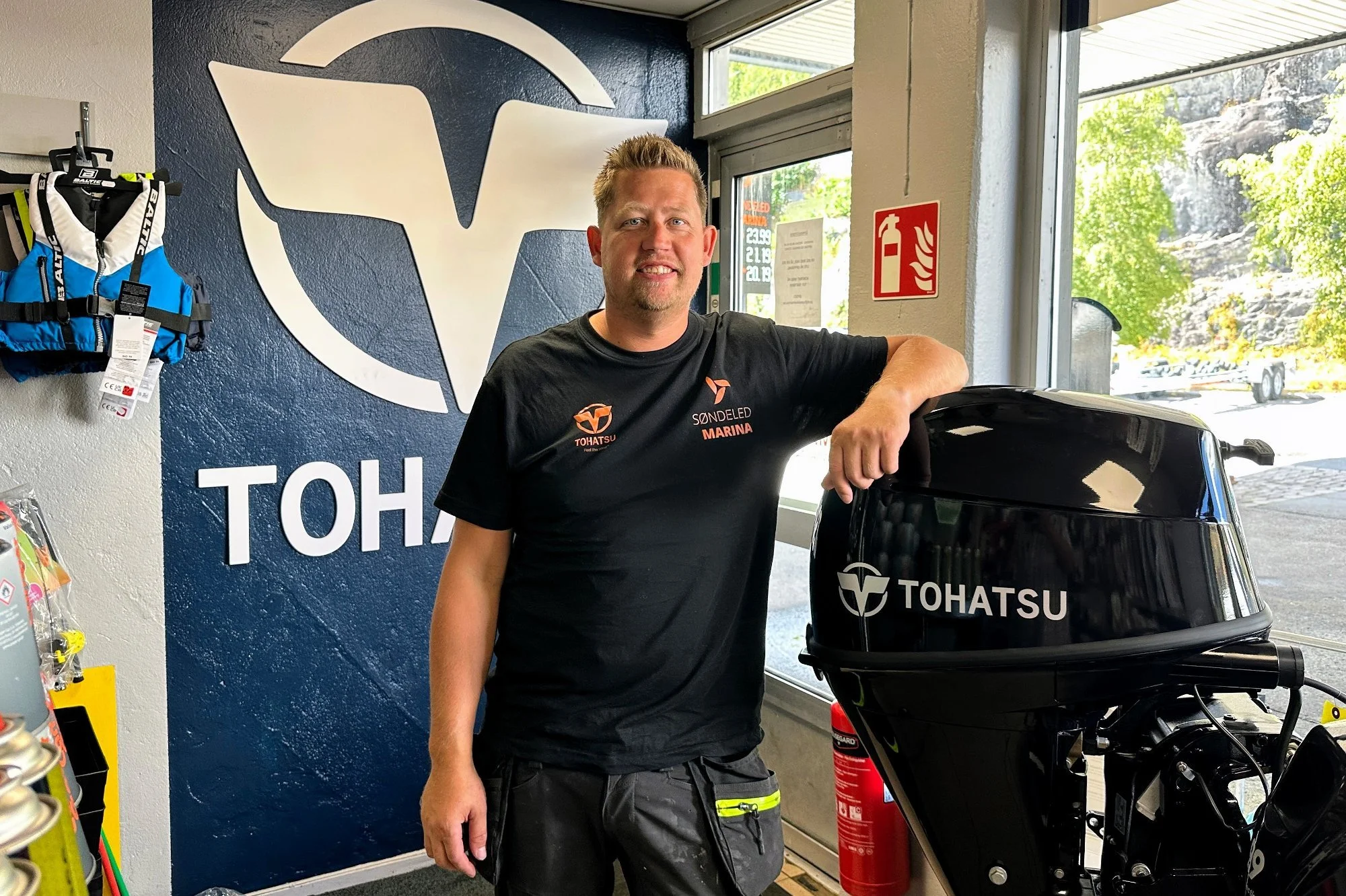 A man with short blond hair and a goatee, wearing a black Tohatsu shirt, standing inside a store next to a Tohatsu outboard motor, with a large blue Tohatsu logo on the wall behind him and a window showing green trees outside.