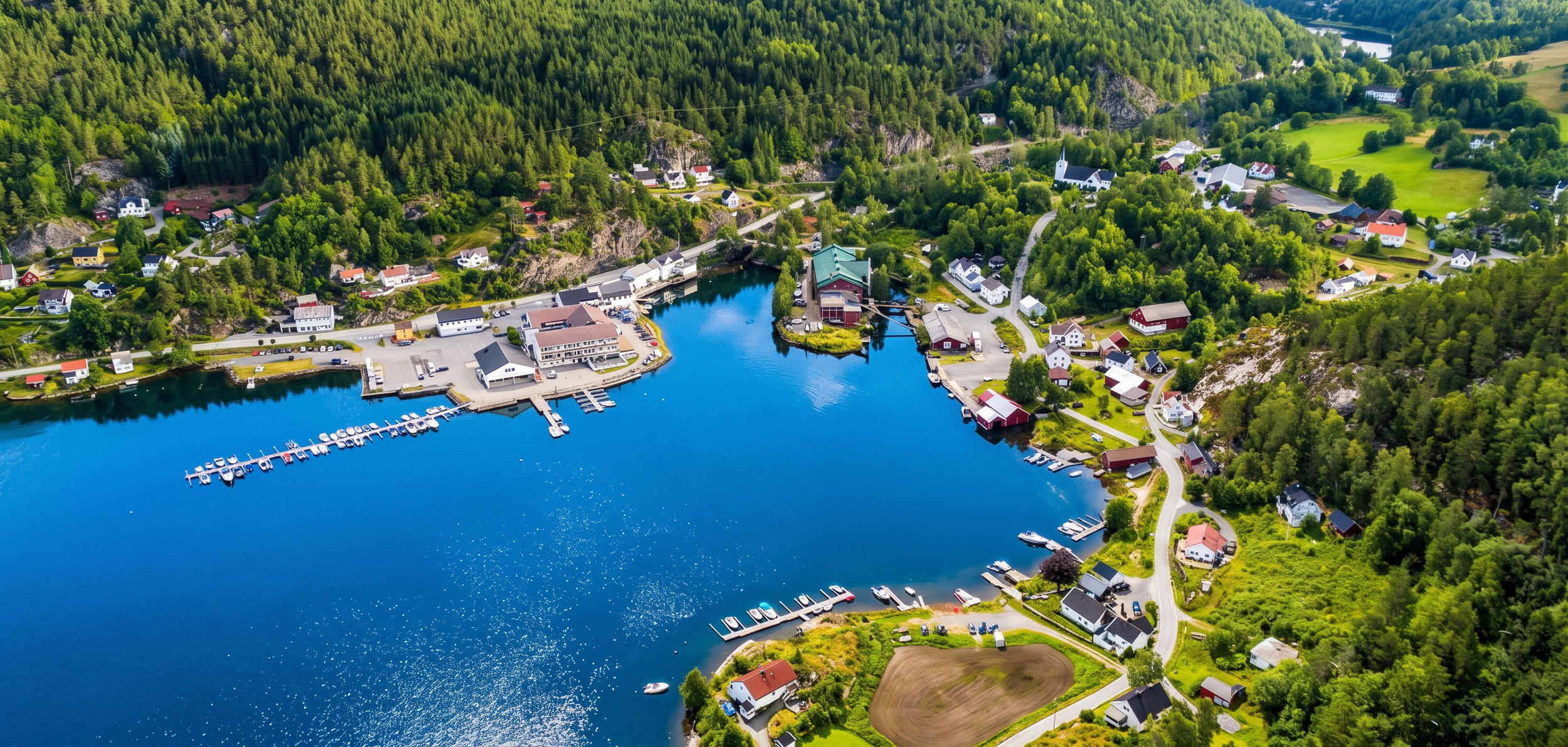 Aerial view of a small harbor with boats, surrounded by green forested hills and scattered houses and buildings.