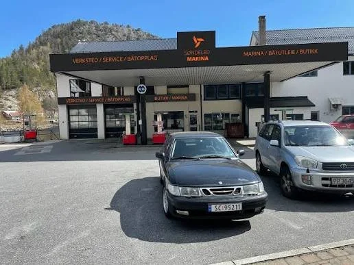 Photo of a marina with a service and boat filling station, parking lot with three cars, and a building with signage in Norwegian and English.